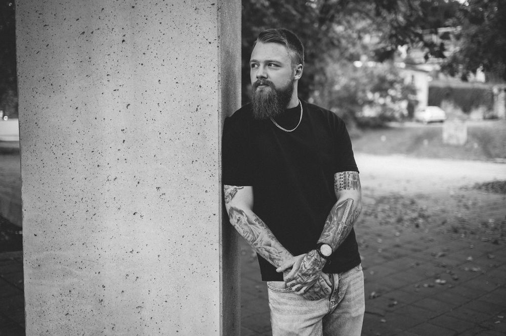 Bearded man with tattooed arms and a chain necklace leans against a concrete pillar outdoors in a black and white portrait.