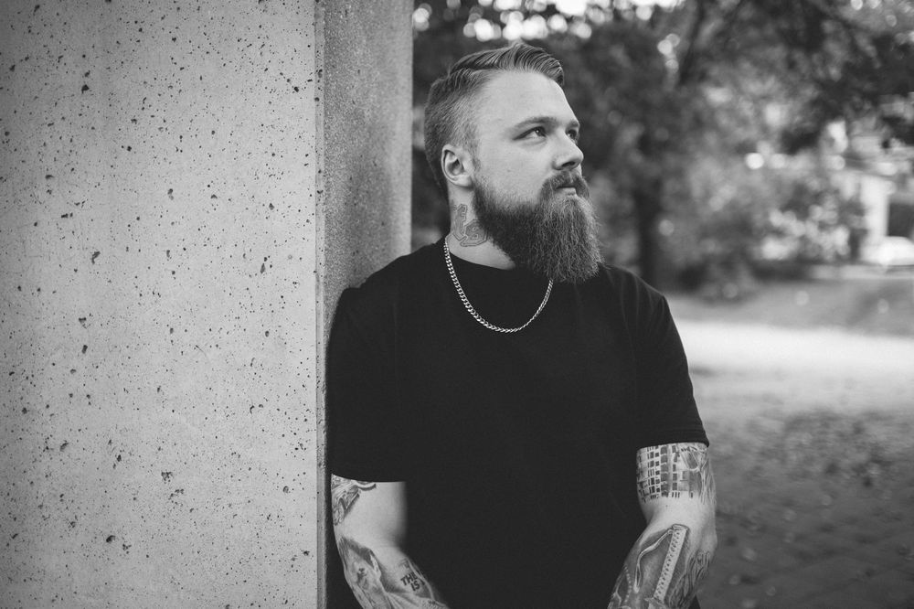 Black and white portrait of a bearded man with tattooed arms and a chain necklace leaning against a concrete pillar outdoors.