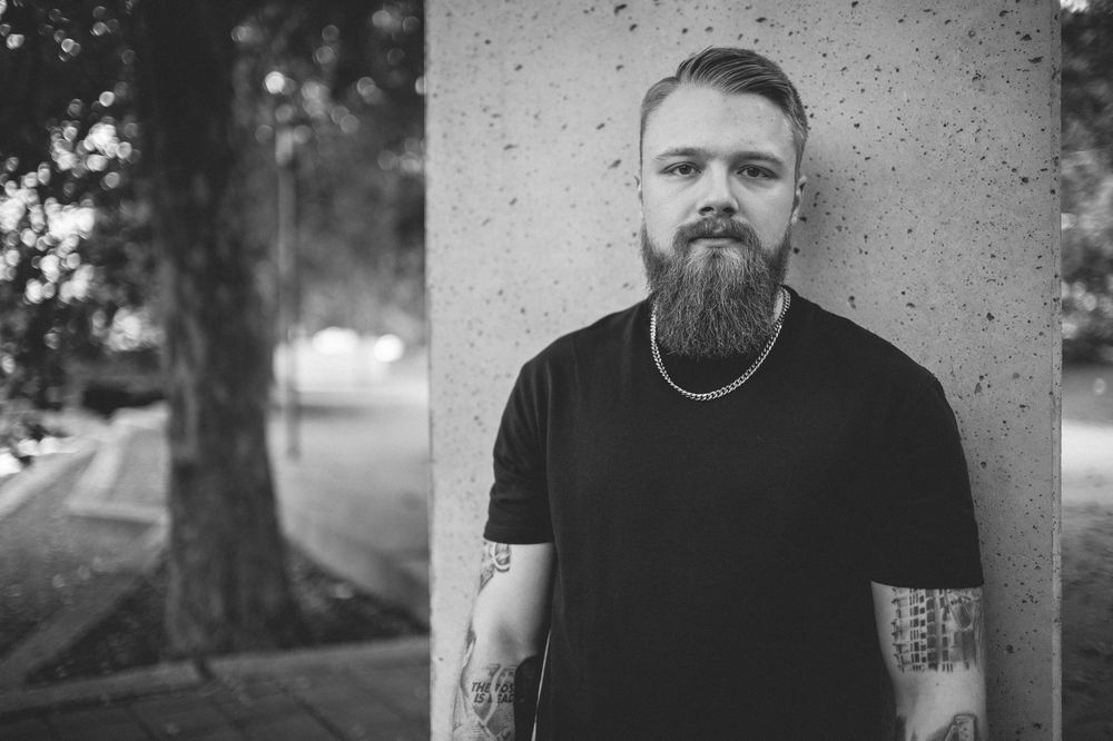 Black and white portrait of a bearded man with tattooed arms and a chain necklace leaning against a concrete pillar outdoors.