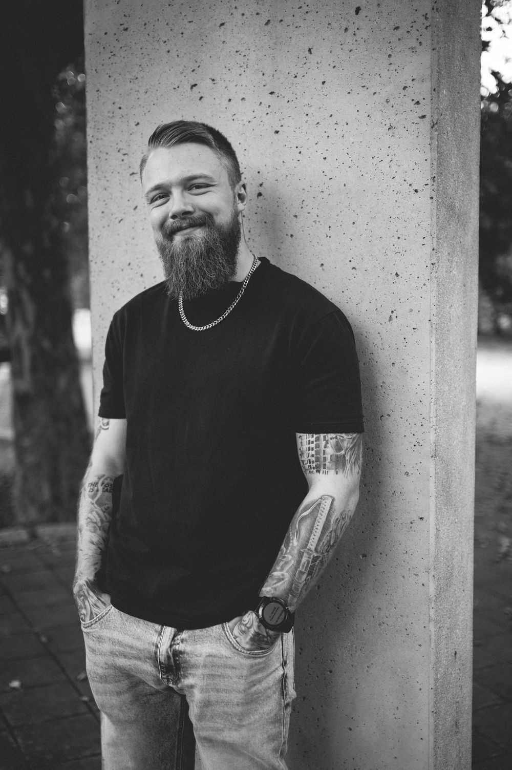 Smiling bearded man with tattoos and a chain necklace leans against a concrete pillar in a black and white photo.