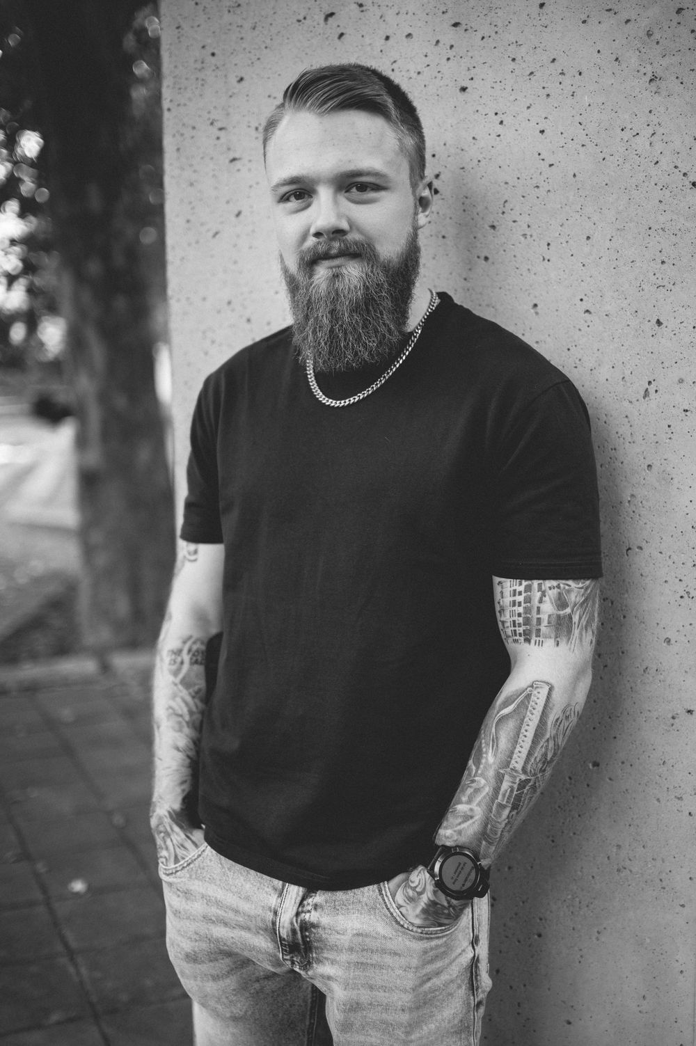 Bearded man with tattooed forearms and a silver chain stands against a concrete wall in a black and white portrait.