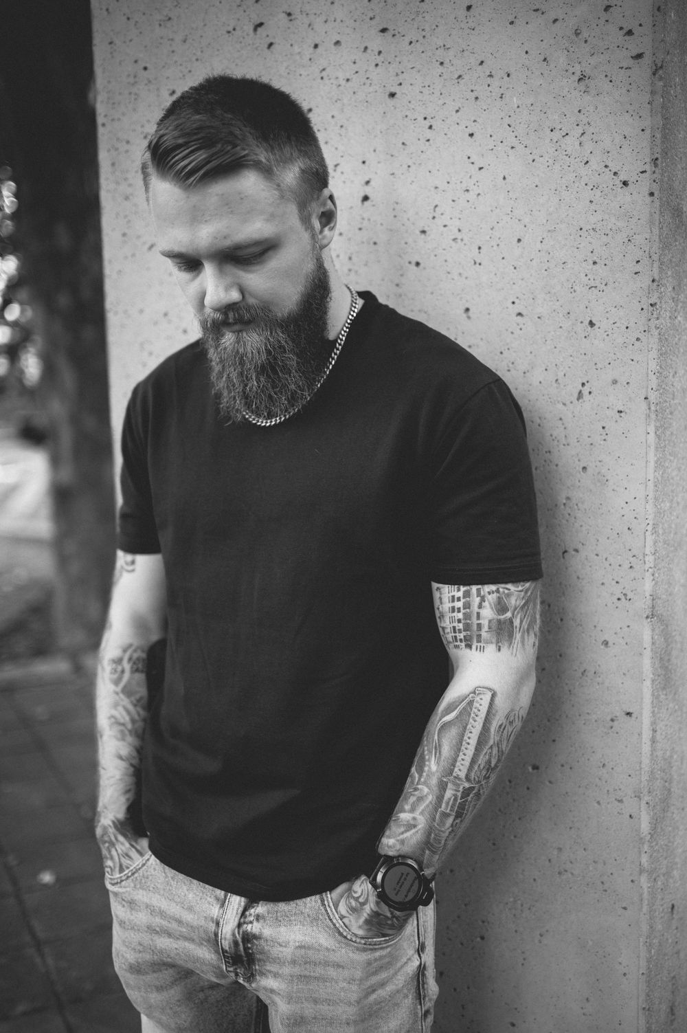 Bearded man with tattooed forearms and a chain necklace leans against a concrete wall, looking down in a black and white portrait.