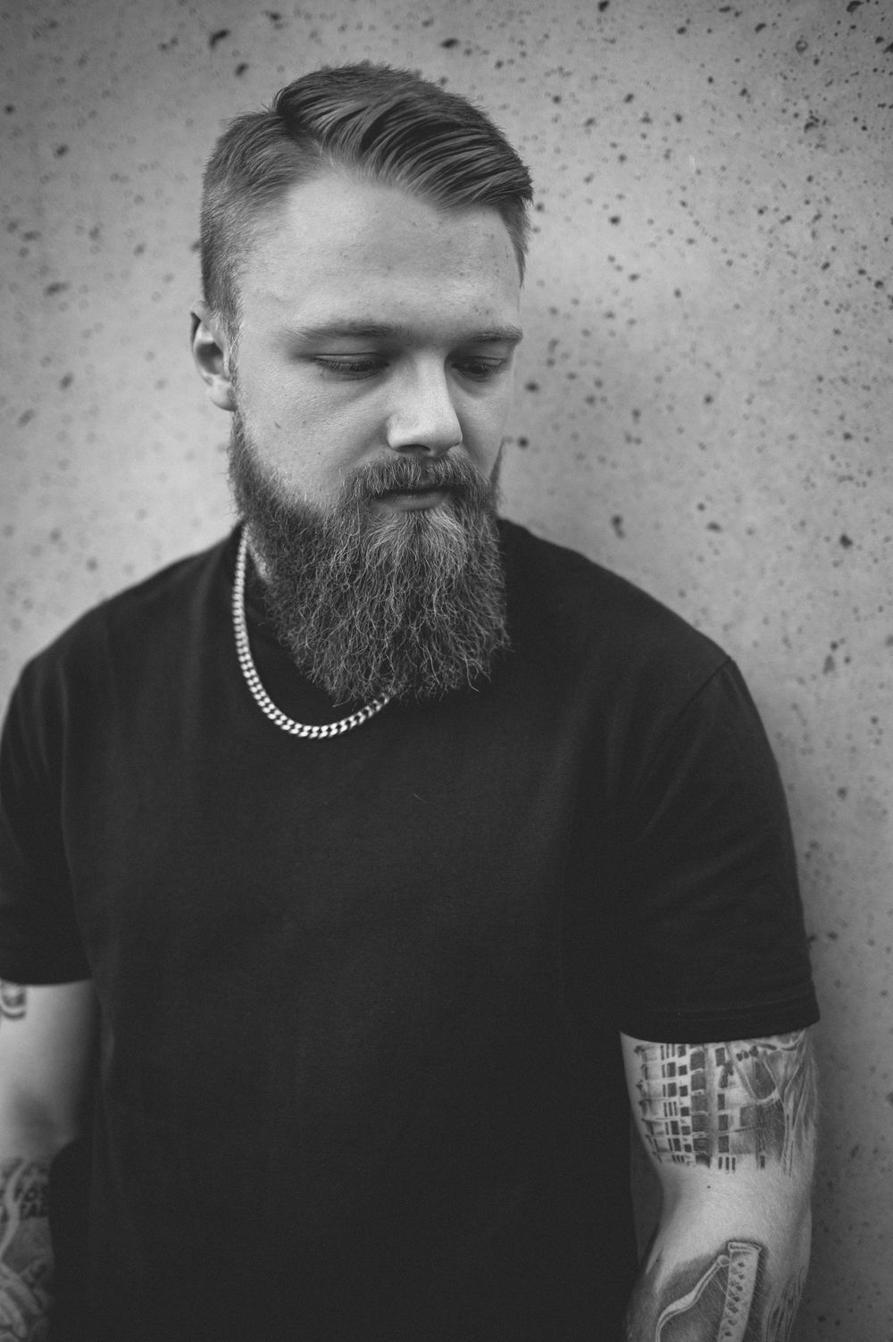 Black and white portrait of a bearded man with tattooed forearms and a chain necklace, looking down while leaning against a textured wall.