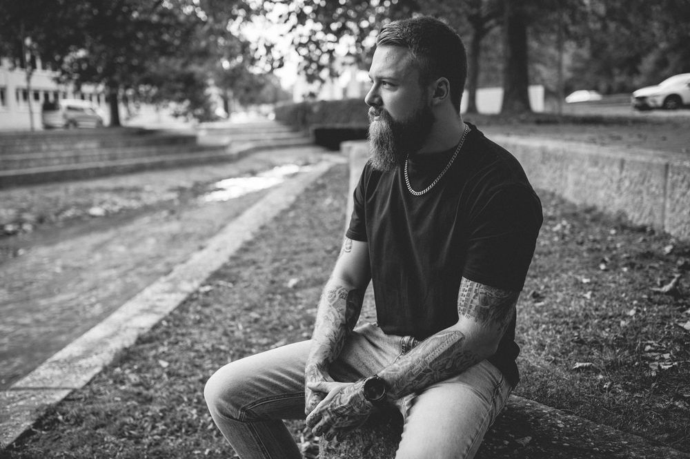 Bearded man with tattooed arms in a dark t-shirt and chain necklace sits outdoors on a stone ledge in a park, shown in black and white.