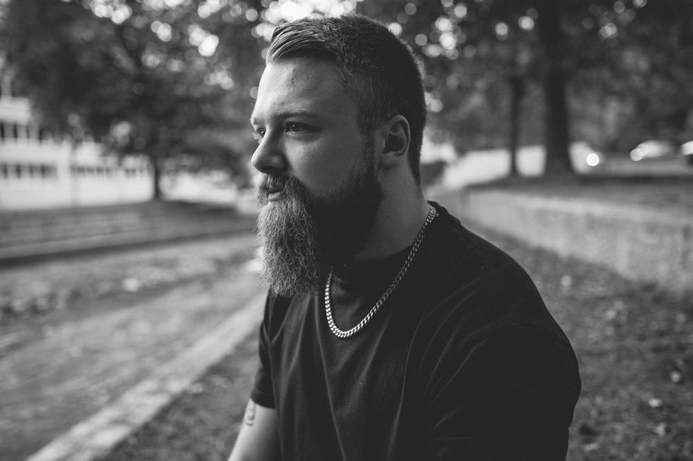 Black and white side profile of a bearded man in a black t-shirt and chain necklace sitting outdoors near trees.