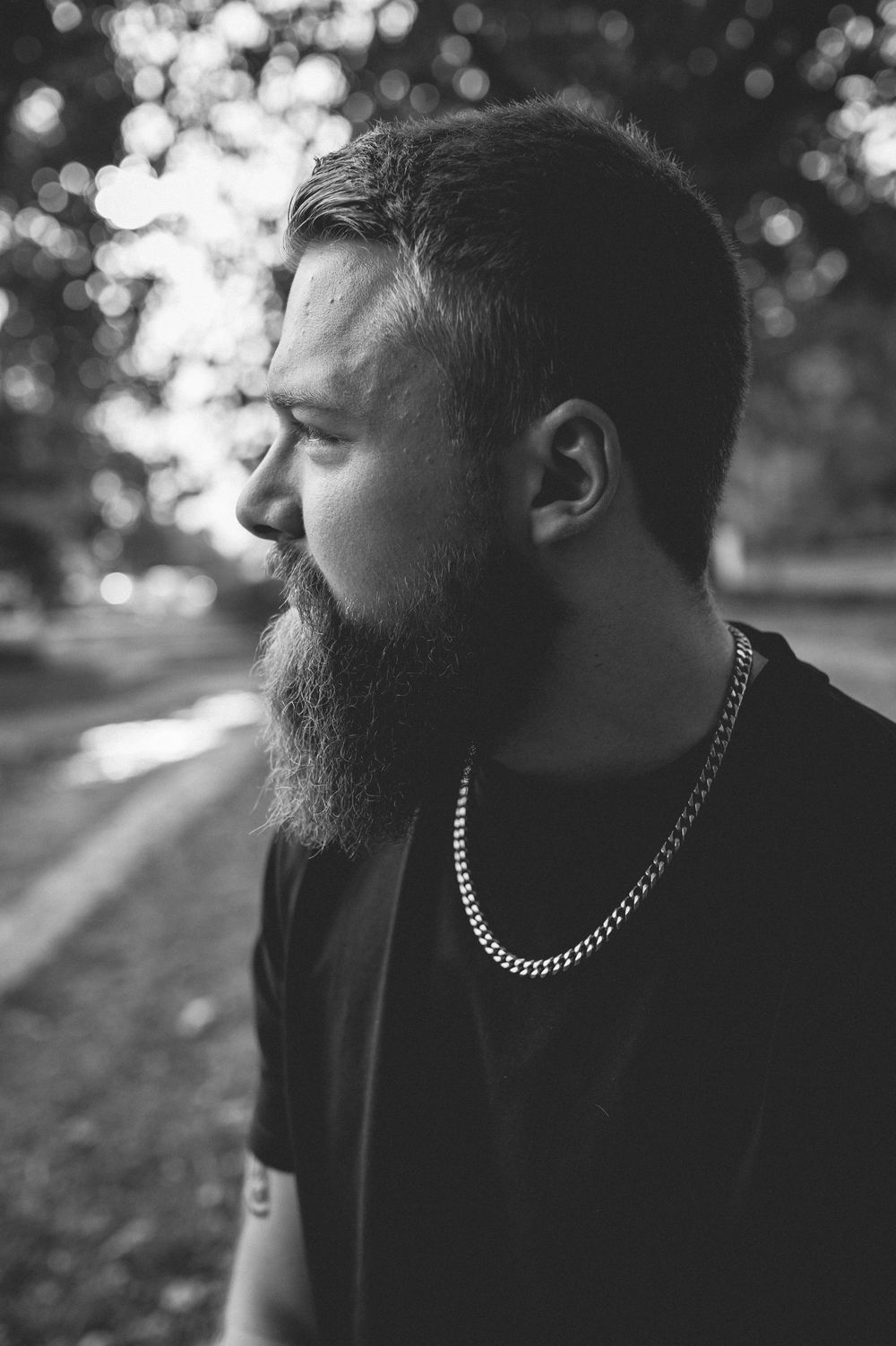 Black and white side profile of a bearded man in a dark t-shirt and silver chain necklace, with blurred trees in the background.