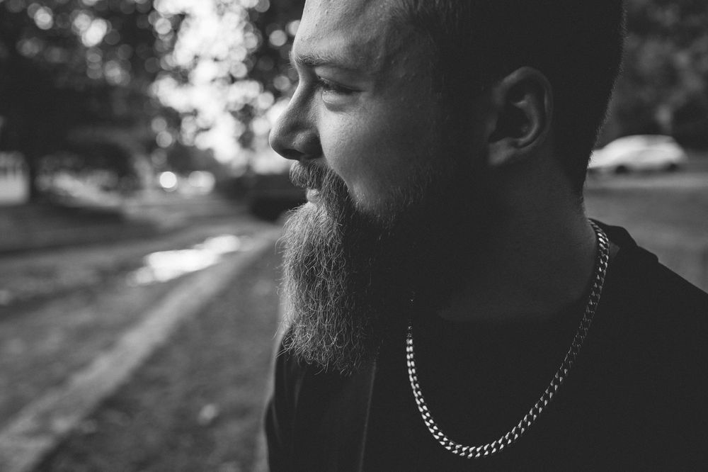 Close-up black and white profile of a smiling bearded man wearing a chain necklace, with a blurred park path and parked car behind him.