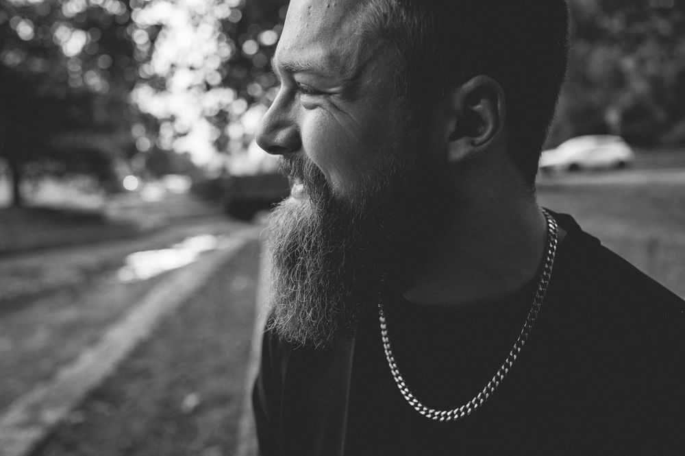 Black and white profile of a smiling bearded man in a dark shirt and chain necklace, standing outdoors near a path with blurred trees.