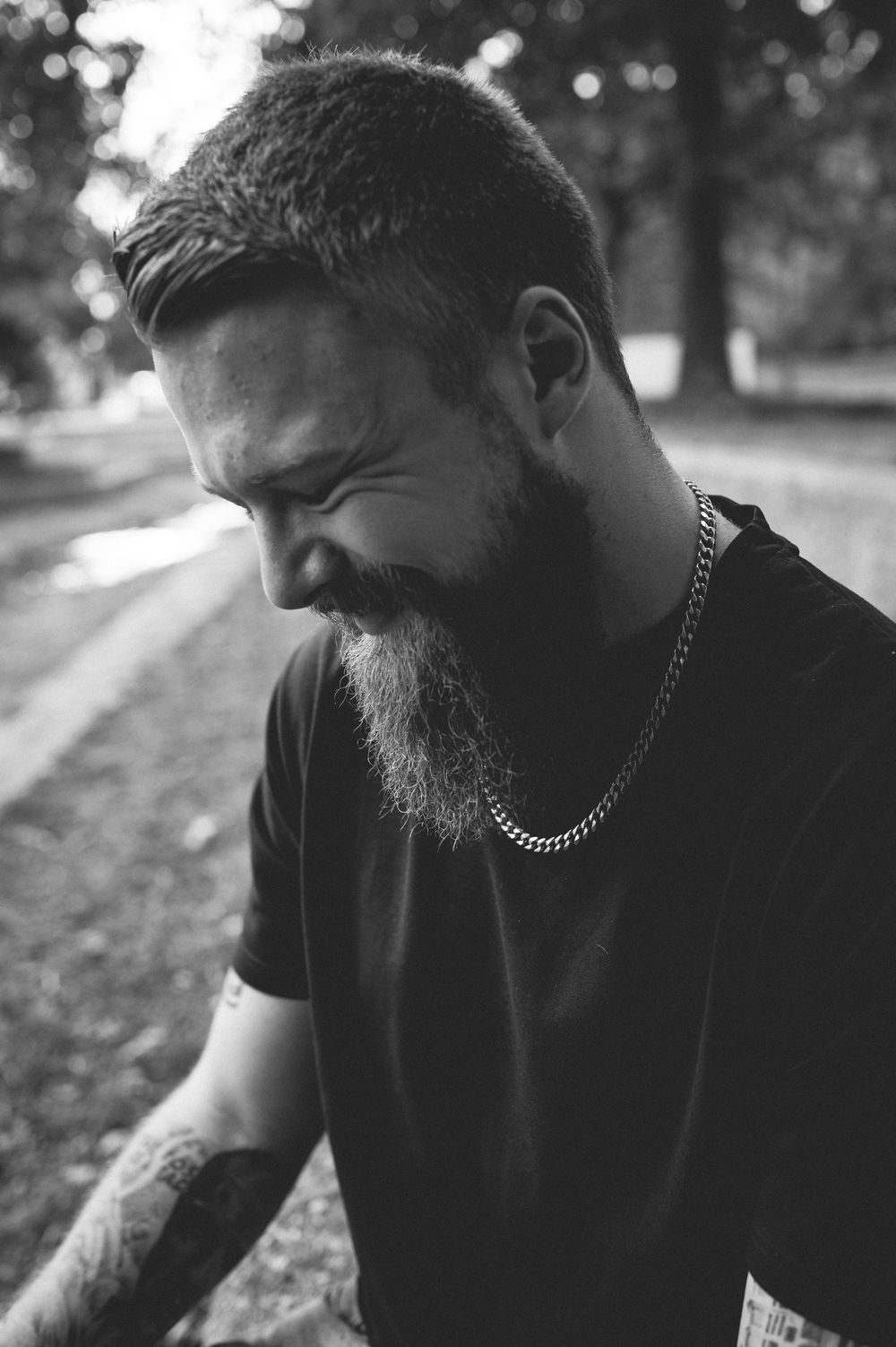 Black and white portrait of a smiling bearded man with a silver chain necklace and arm tattoos looking down in an outdoor park setting.