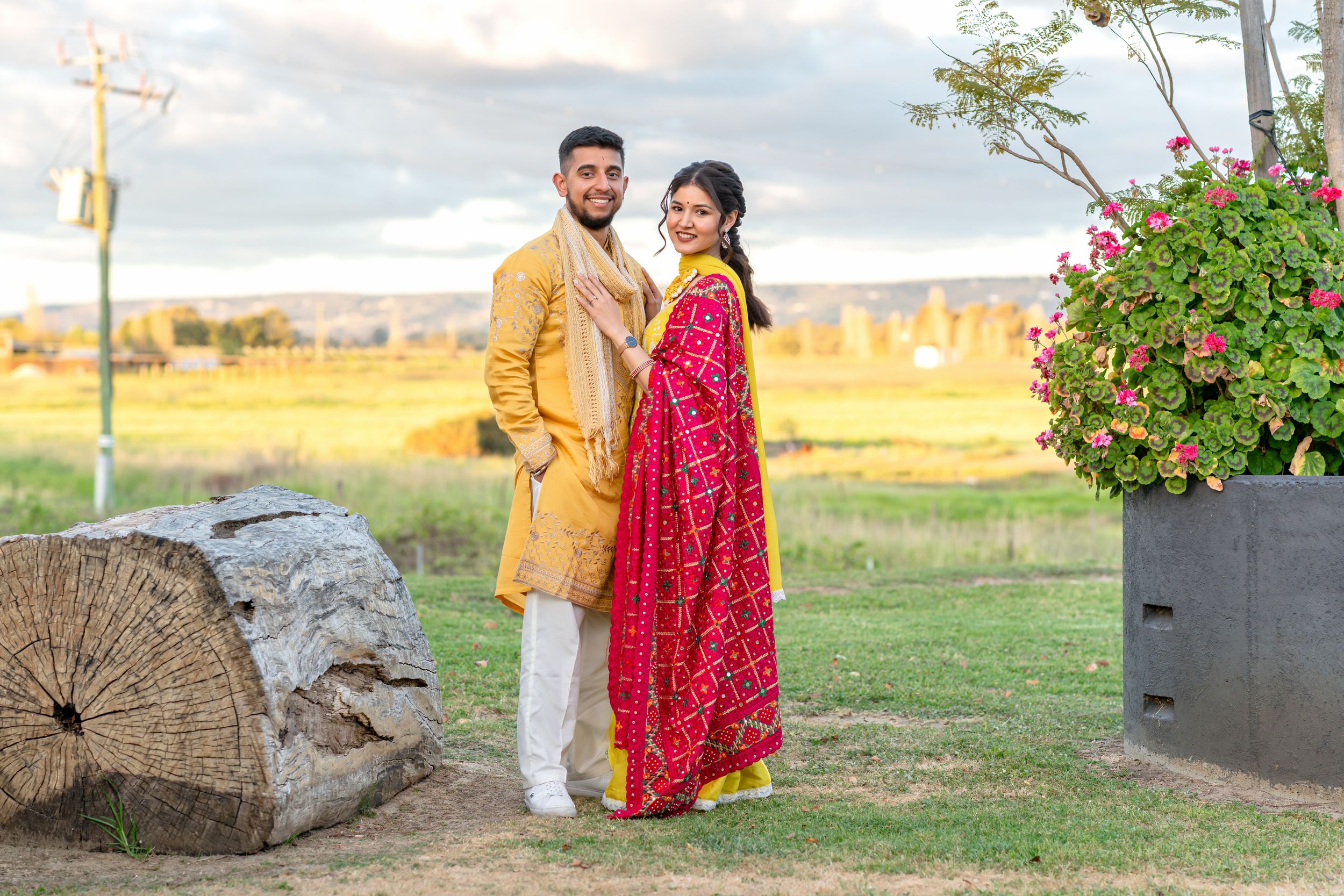Couple in traditional South Asian attire posing outdoors, man in gold sherwani and woman in vibrant red saree.