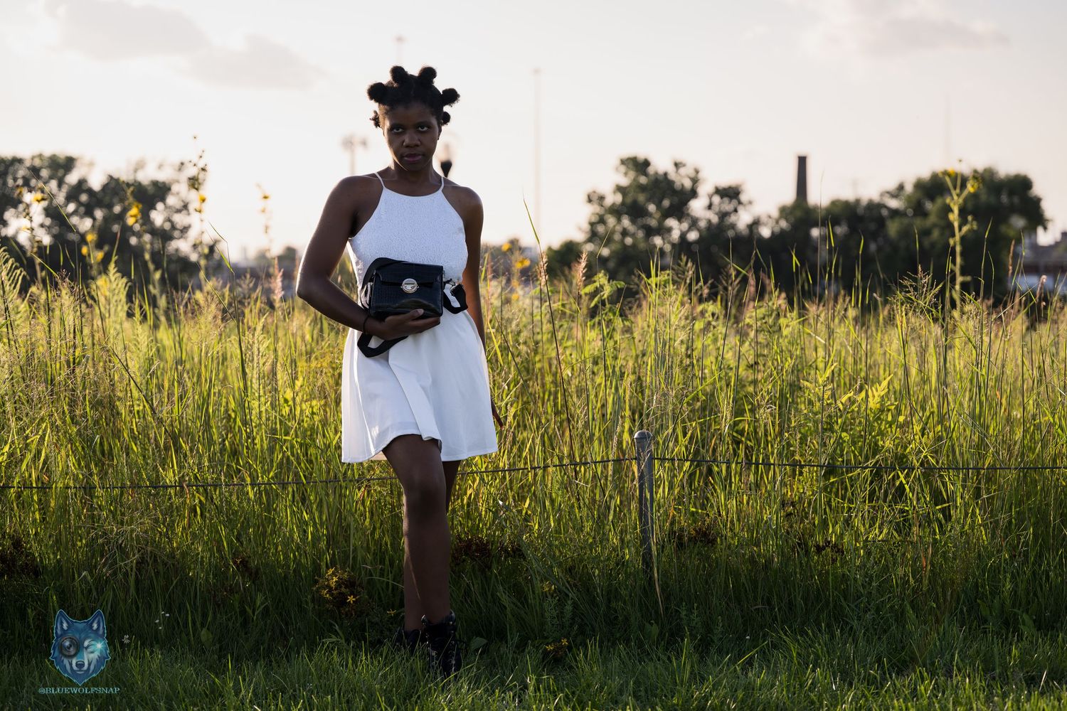 A young Black woman in a white dress holds a clutch purse while standing in a lush green field at golden hour.