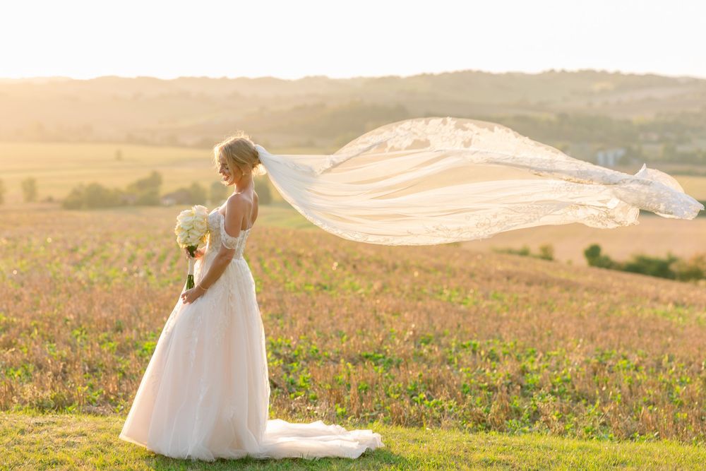 Bride in white gown holds bouquet as veil flows in wind across golden sunlit field.
