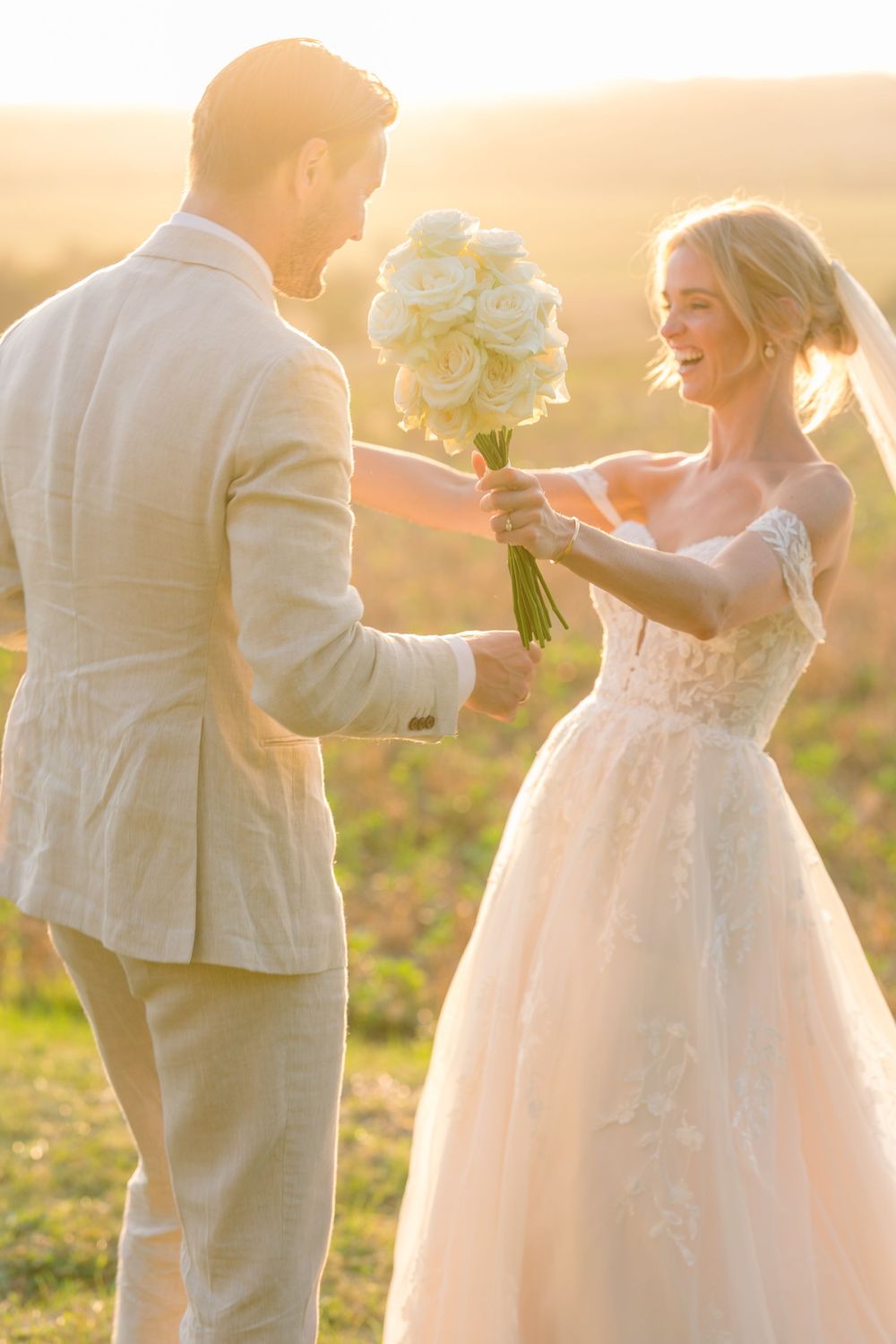 A smiling bride in lace gown holds white roses bouquet with groom in beige suit at golden sunset.