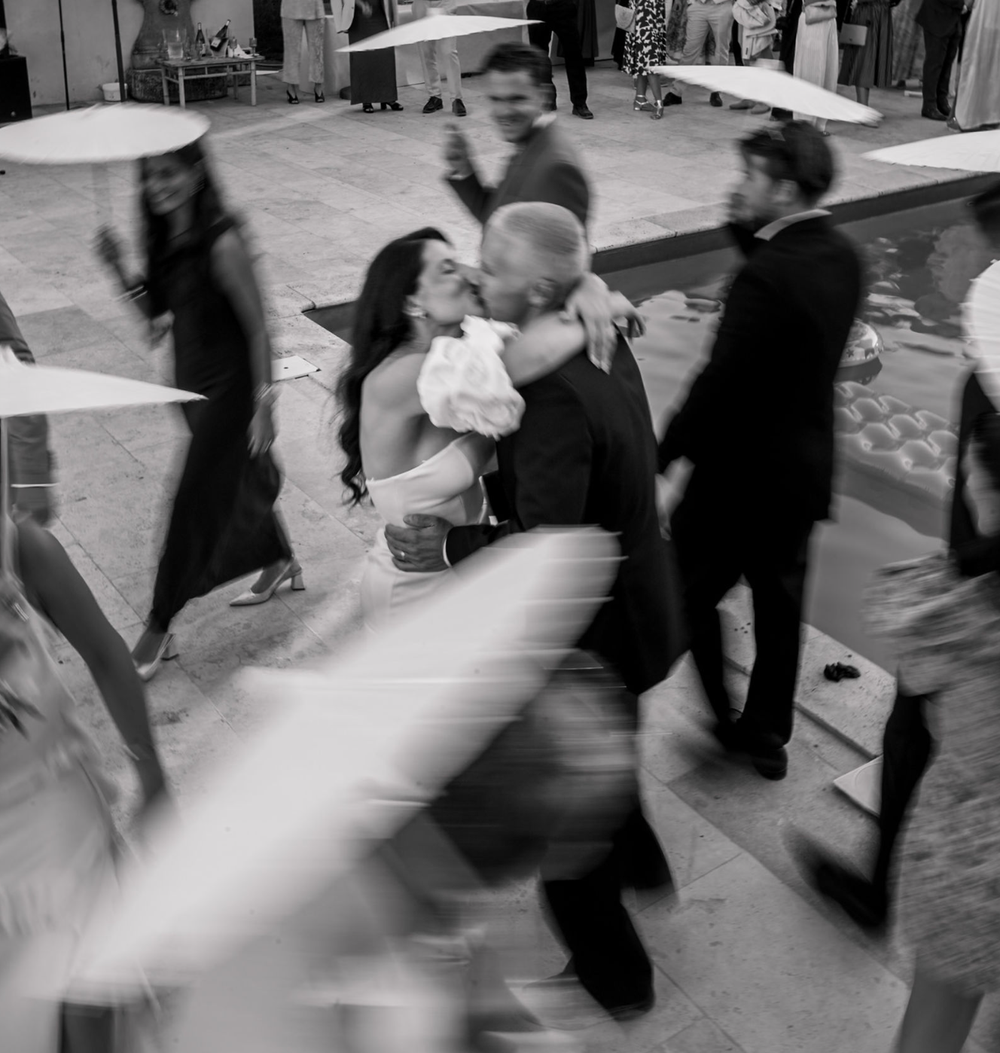 Black and white wedding photo of a bride and groom kissing as blurred guests dance and walk around them by a pool.
