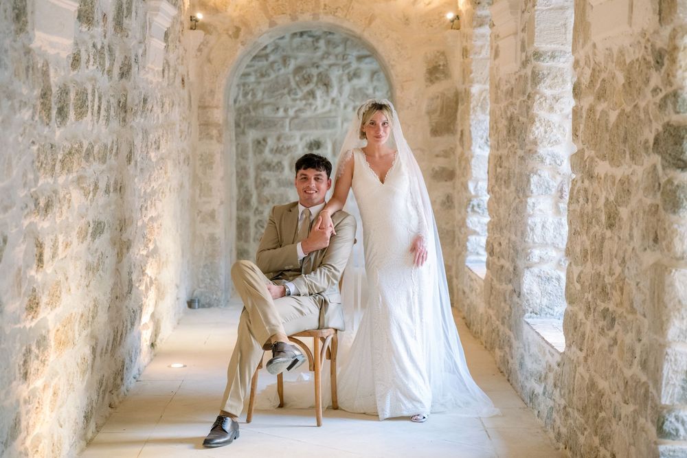 Bride and groom posing in a rustic stone archway corridor, bride in white gown with veil, groom seated in chair.