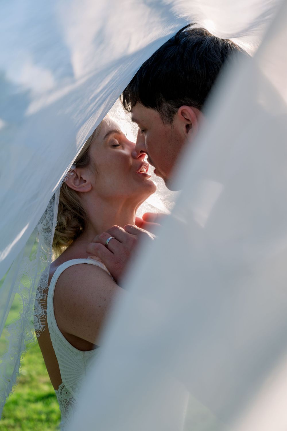 Bride and groom sharing a romantic kiss under a flowing white wedding veil outdoors on their wedding day.