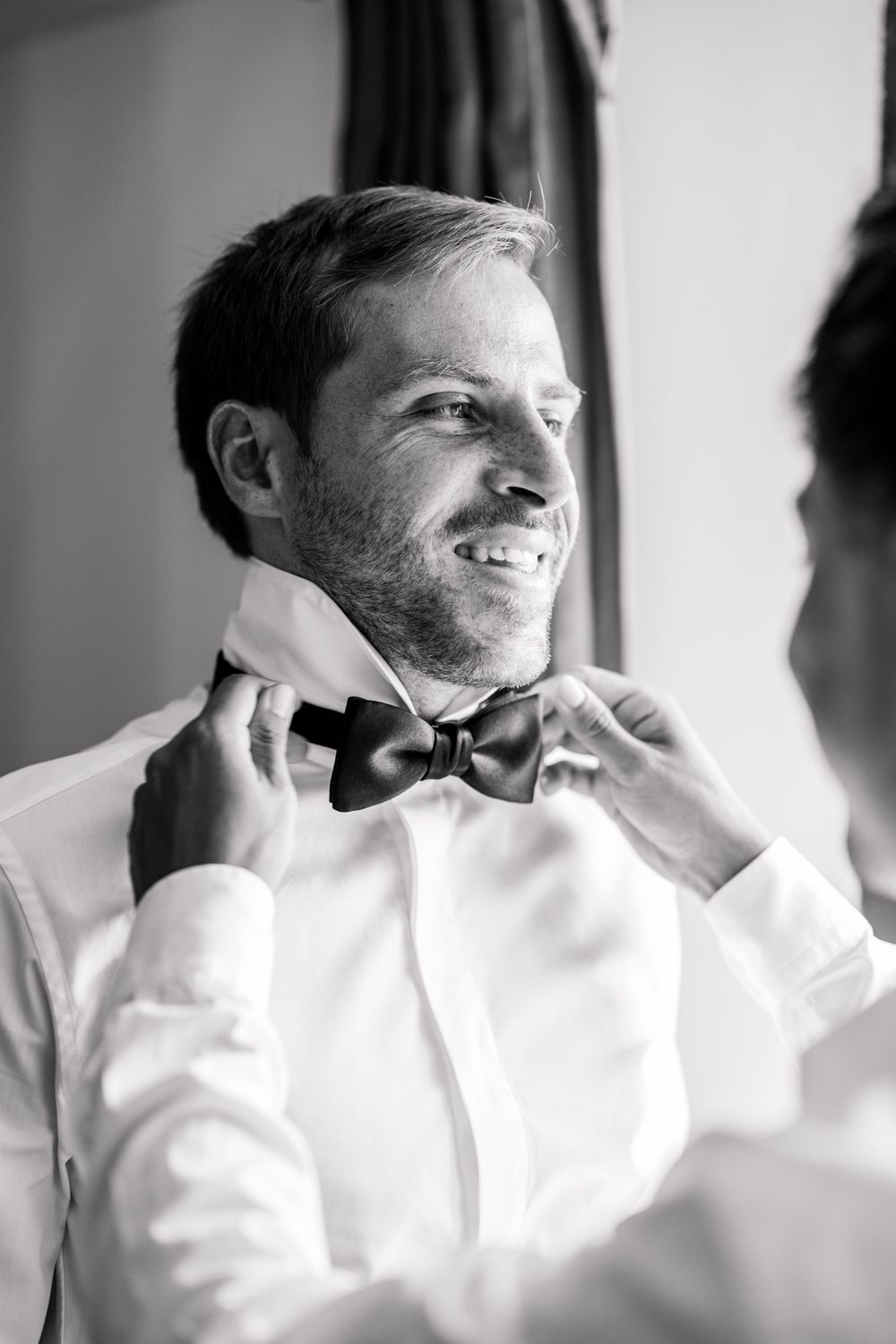 Black and white photo of a smiling groom having his bow tie adjusted by another person while getting ready.
