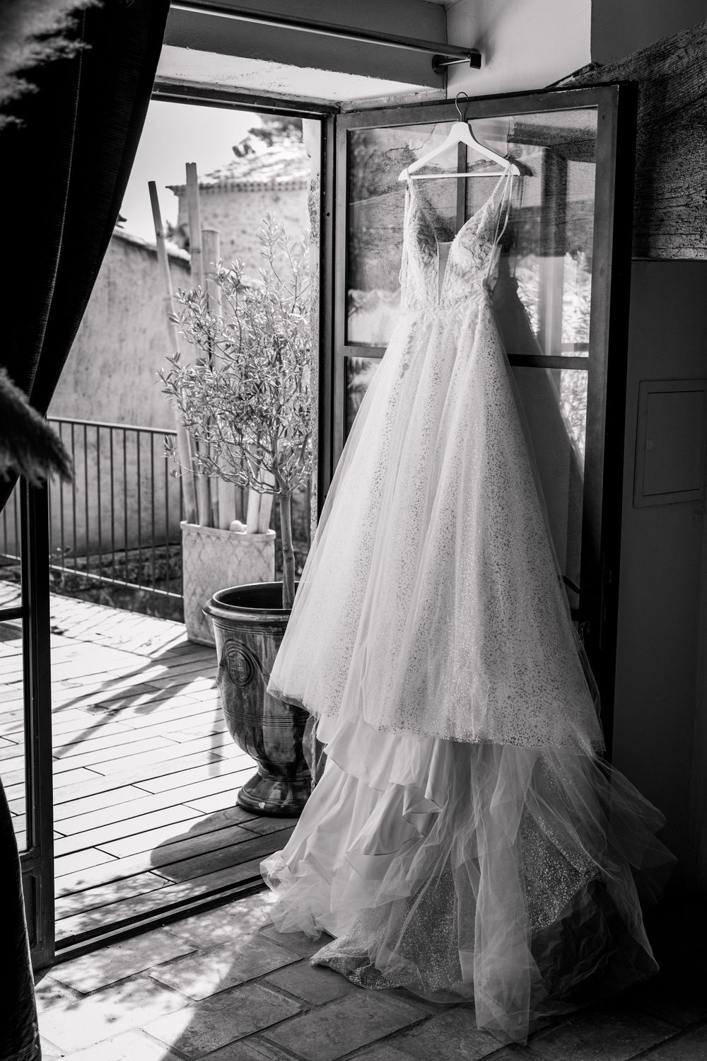 Black and white photo of an elegant lace wedding dress hanging on a hanger in a doorway leading to a sunlit patio.