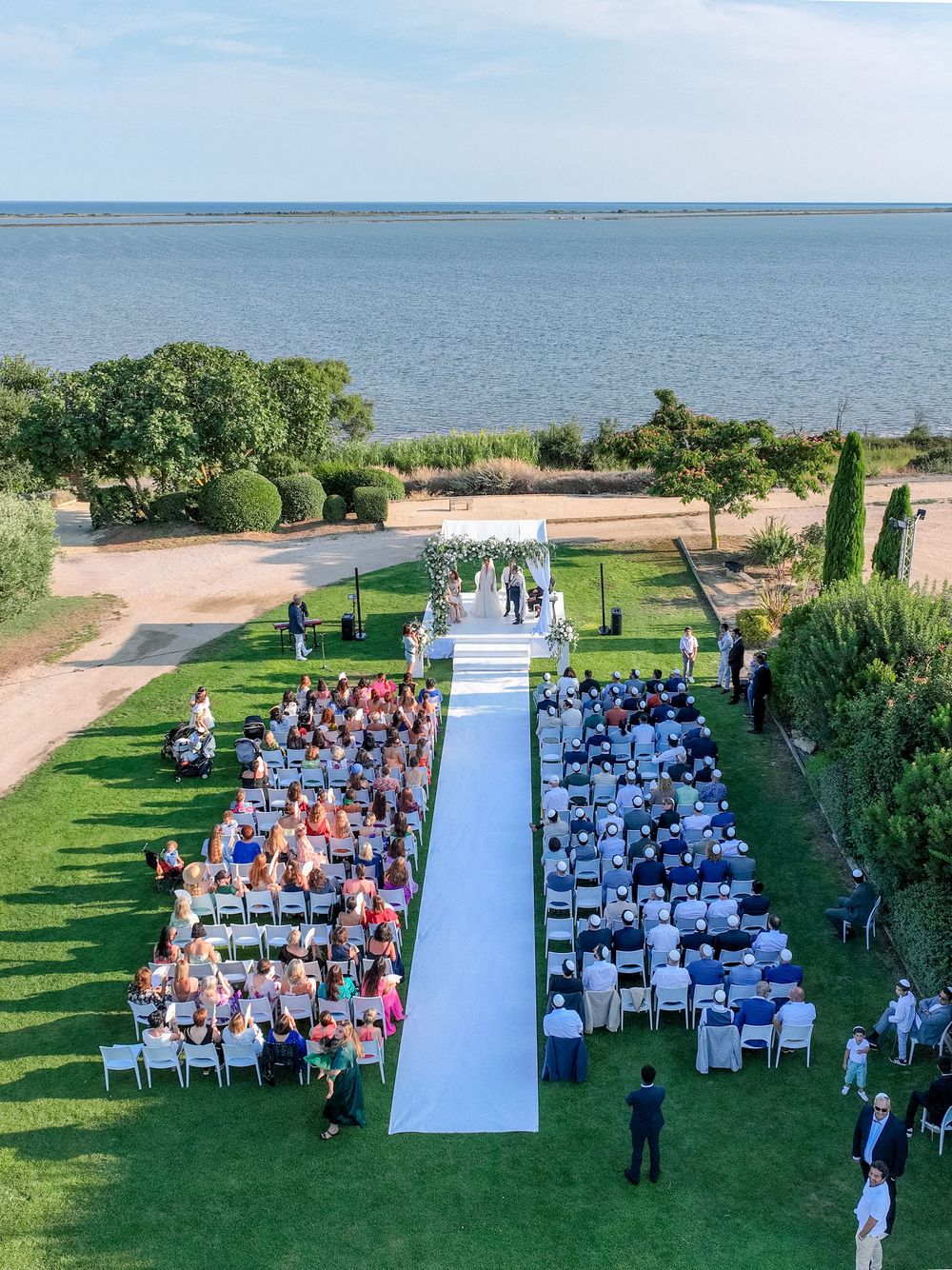 Outdoor wedding ceremony by the sea with white aisle, floral arch, and guests seated on both sides on a lush green lawn.