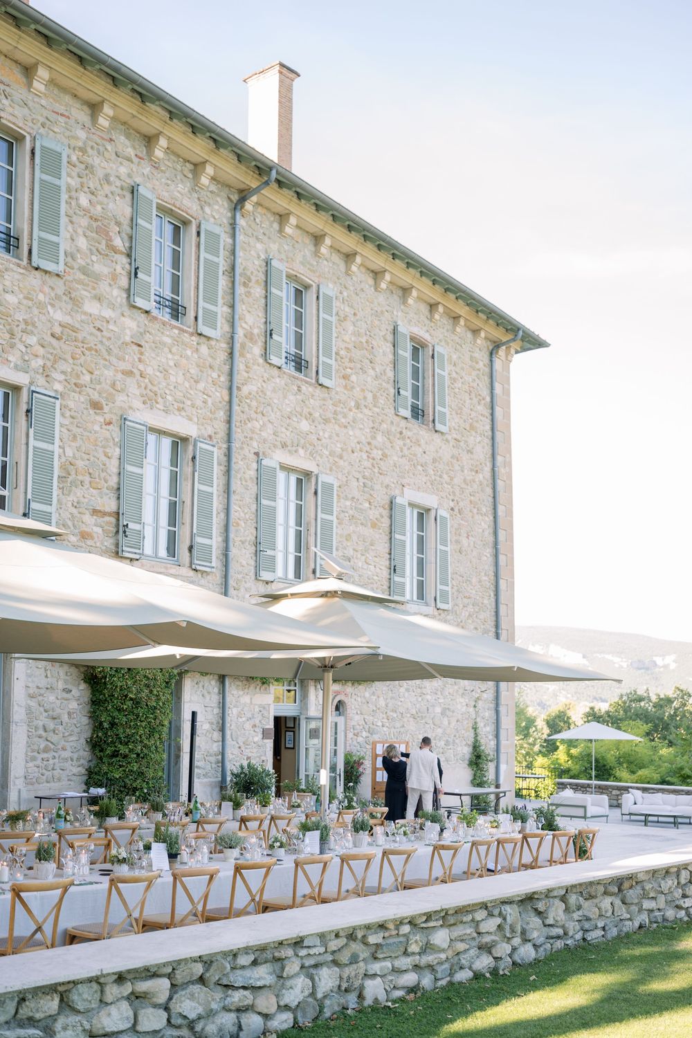Elegant outdoor wedding reception at a stone villa with long tables, white umbrellas, and scenic countryside views.