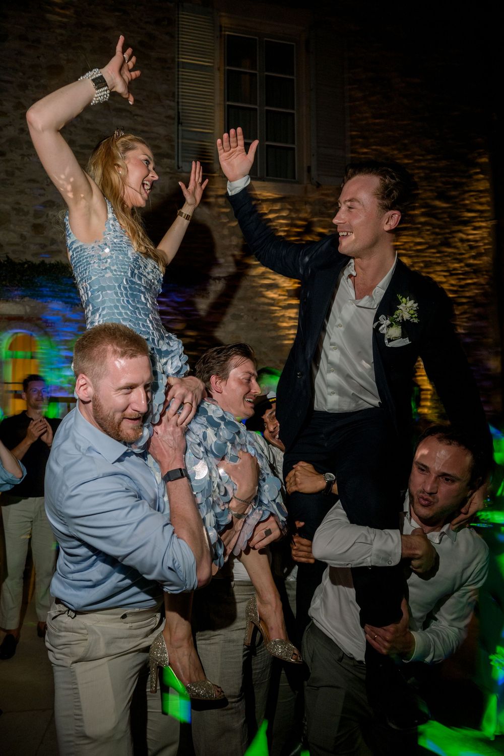 Wedding guests celebrate joyfully as bride and groom are lifted on shoulders during lively outdoor reception dance.
