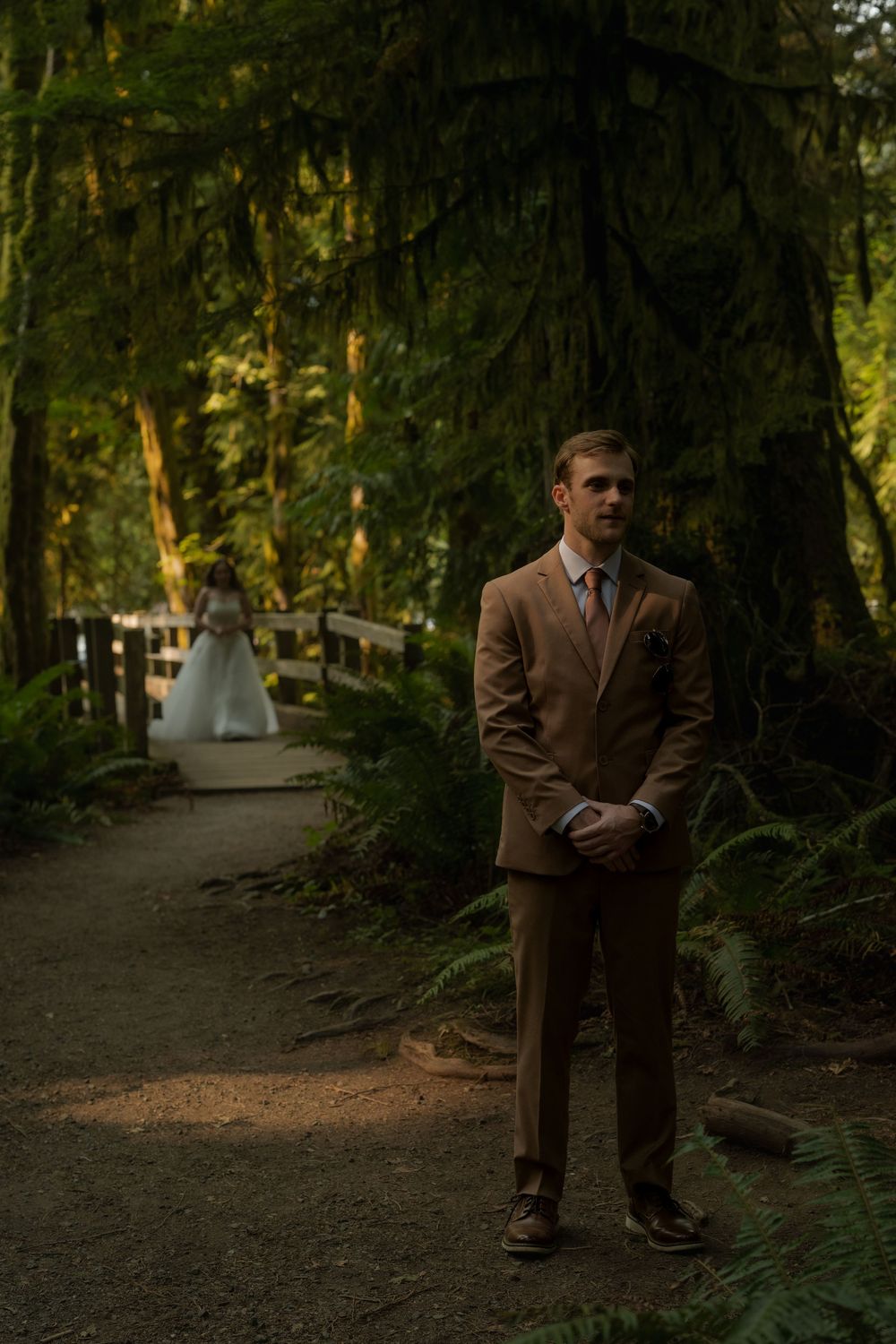 Groom in tan suit stands on forest path with bride approaching in background during outdoor wedding ceremony.