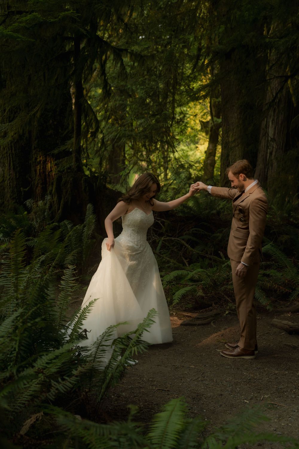 Bride and groom dancing on a forest path surrounded by lush ferns and tall trees.