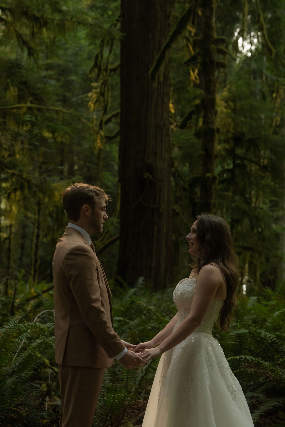 A bride and groom hold hands facing each other in a lush green forest during their intimate woodland wedding ceremony.