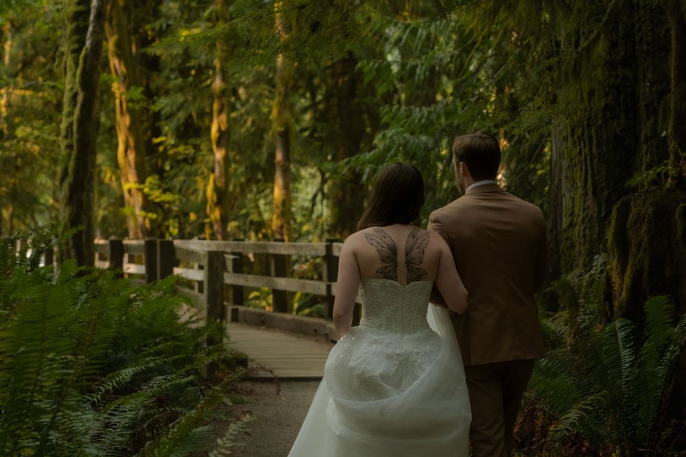 Bride and groom walking hand in hand along a lush forest path, bride wearing a lace-back wedding gown.