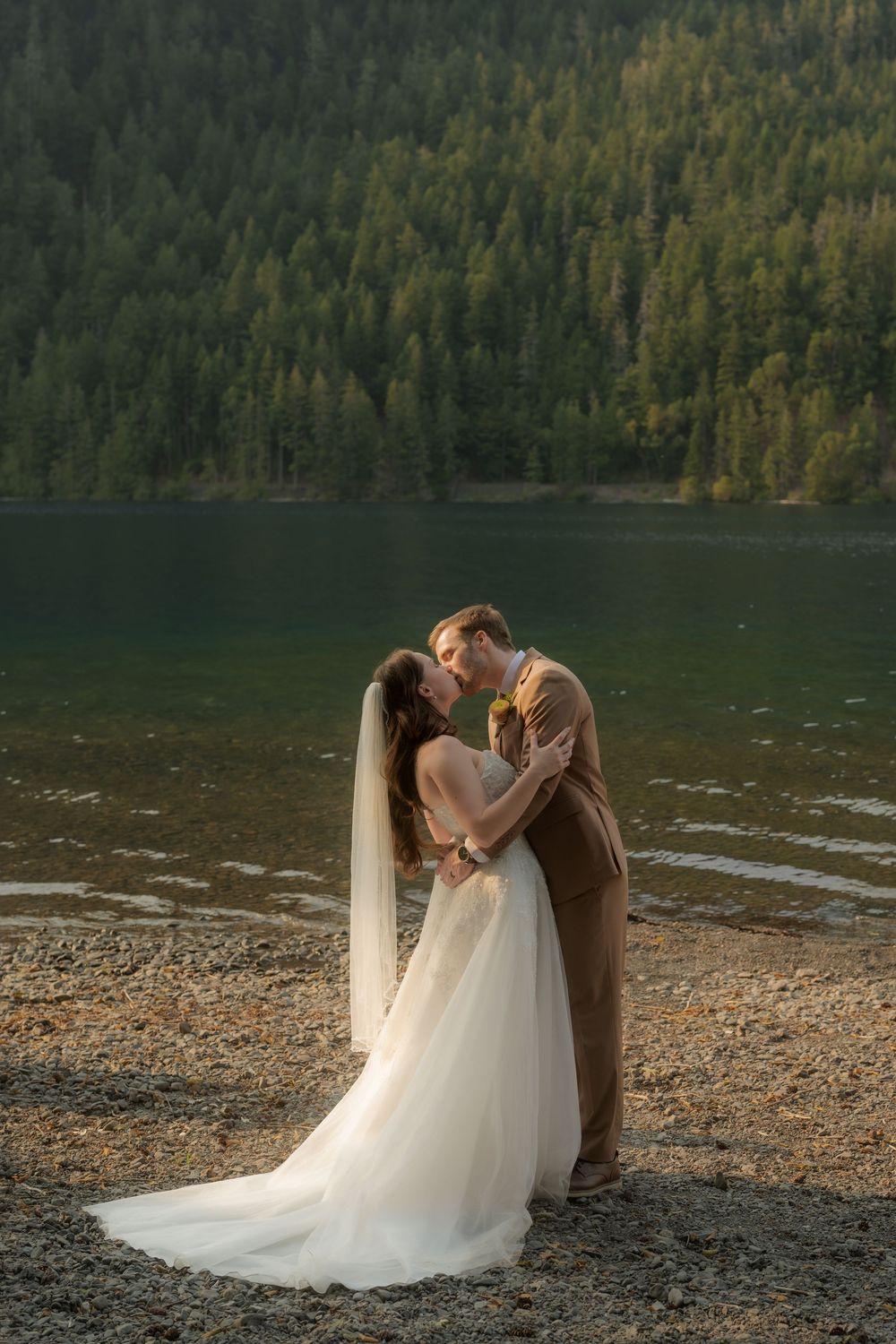 Bride and groom share a romantic kiss by a serene mountain lake surrounded by lush evergreen forest during golden hour.