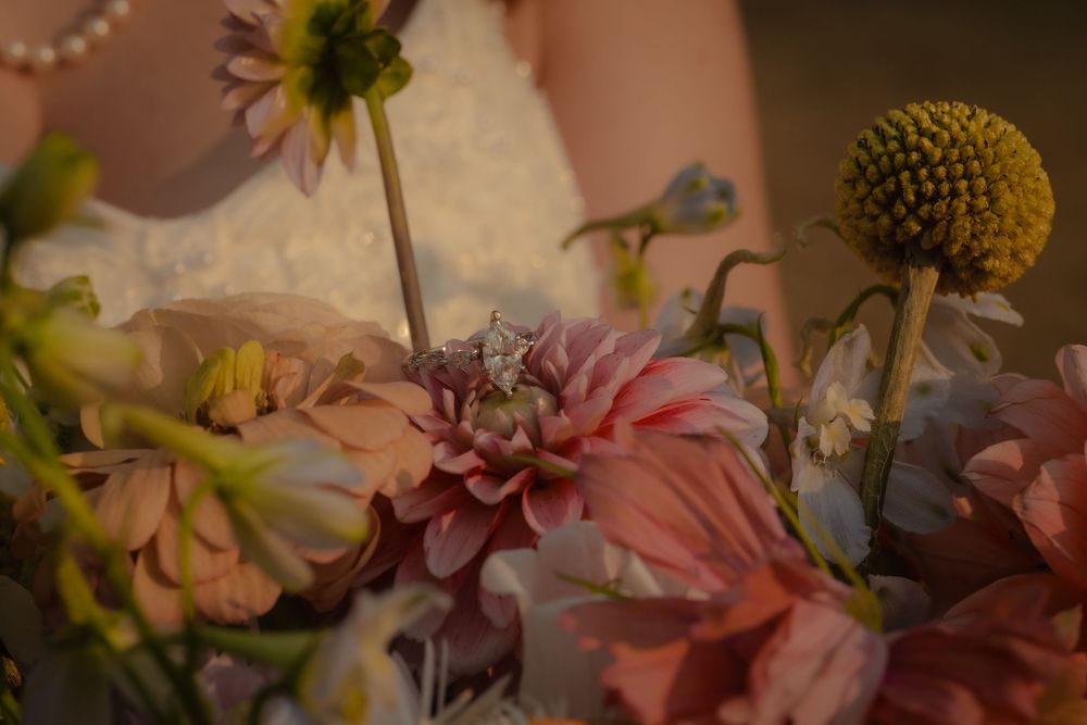Close-up of a diamond engagement ring nestled among pink dahlias and wildflowers in a bridal bouquet.