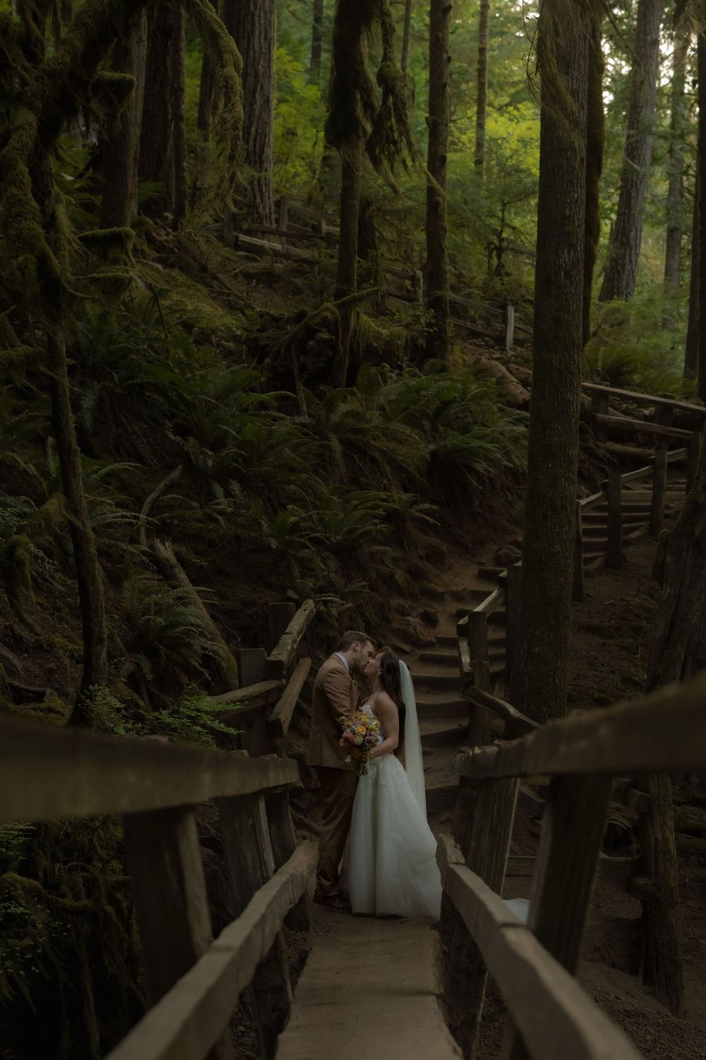 A couple shares a romantic kiss on a wooden staircase surrounded by lush green forest trees.