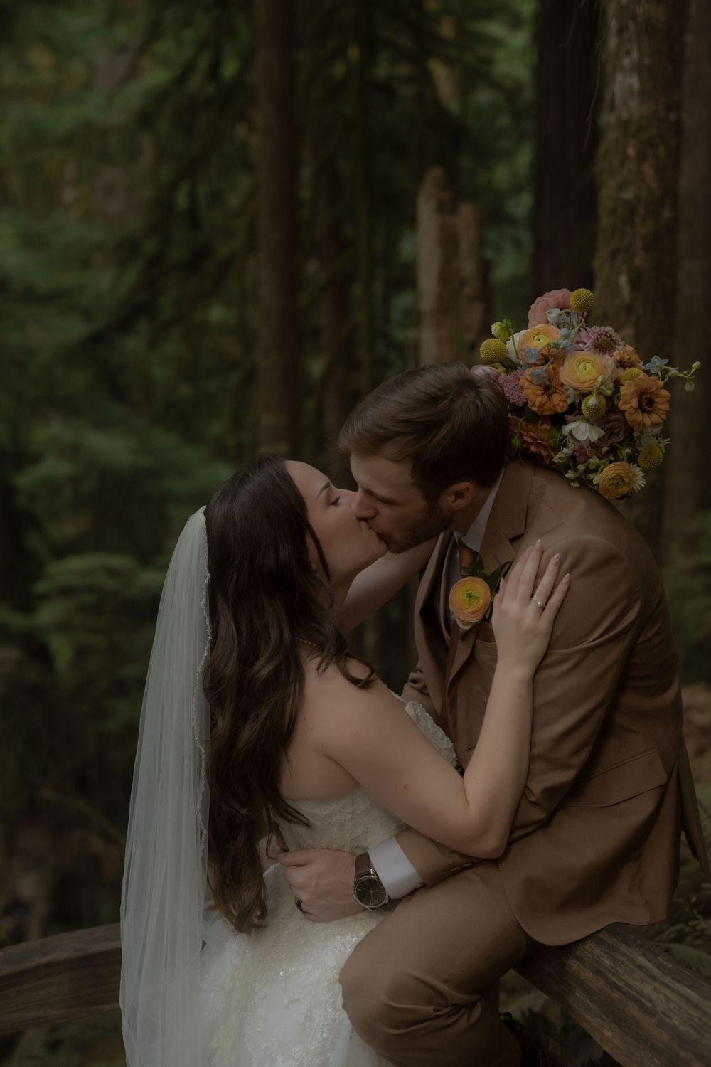 Bride and groom share a romantic kiss in a lush forest setting, with a colorful bouquet and wedding attire.