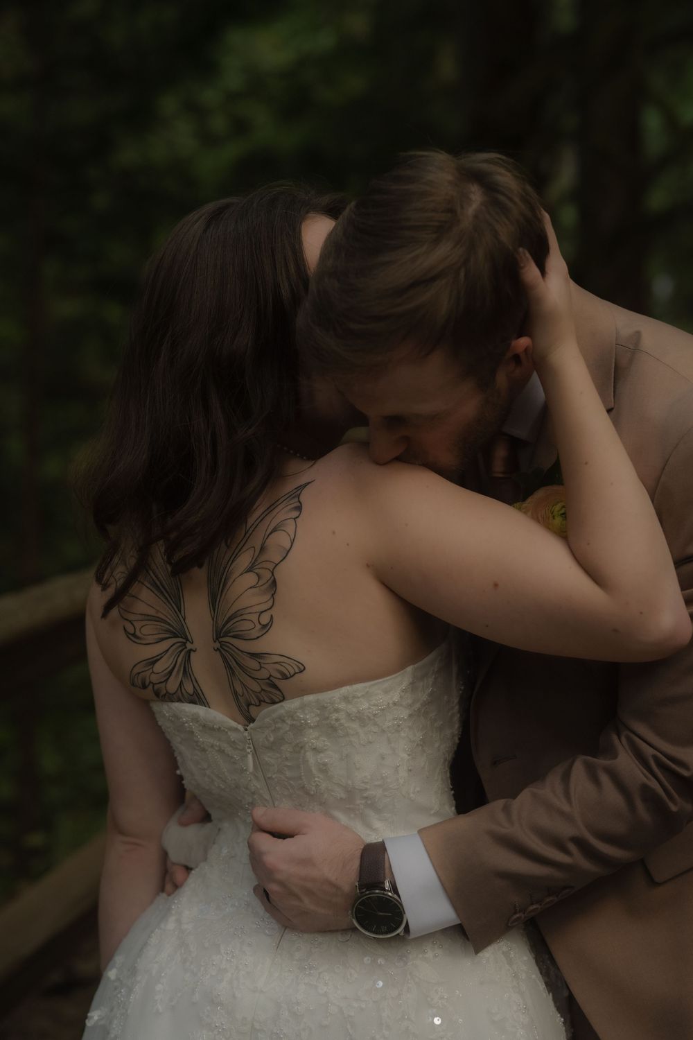 Bride with butterfly back tattoo embracing groom in wedding attire among lush green forest backdrop.