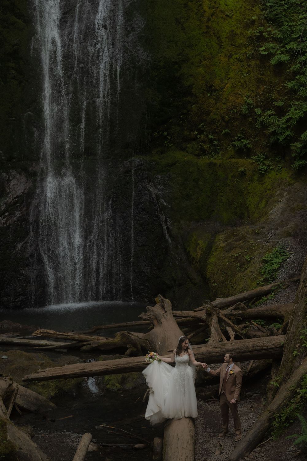 Bride and groom hold hands on logs before a lush waterfall surrounded by moss-covered cliffs.