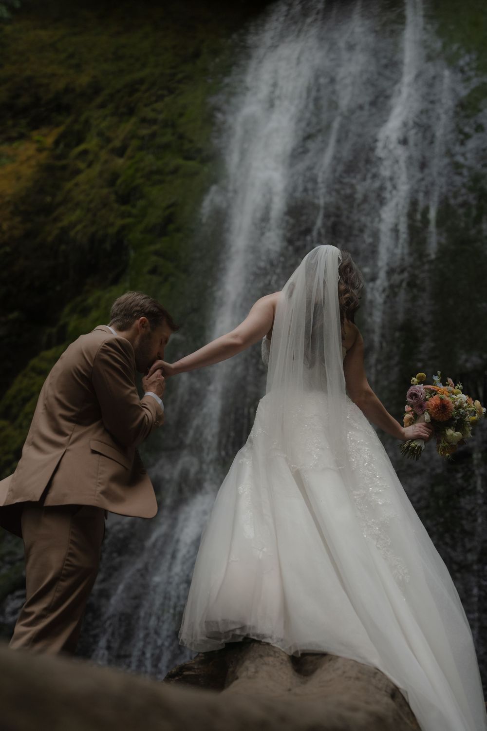 Bride in white gown and veil holds groom's hand near a misty waterfall, holding a colorful bouquet.