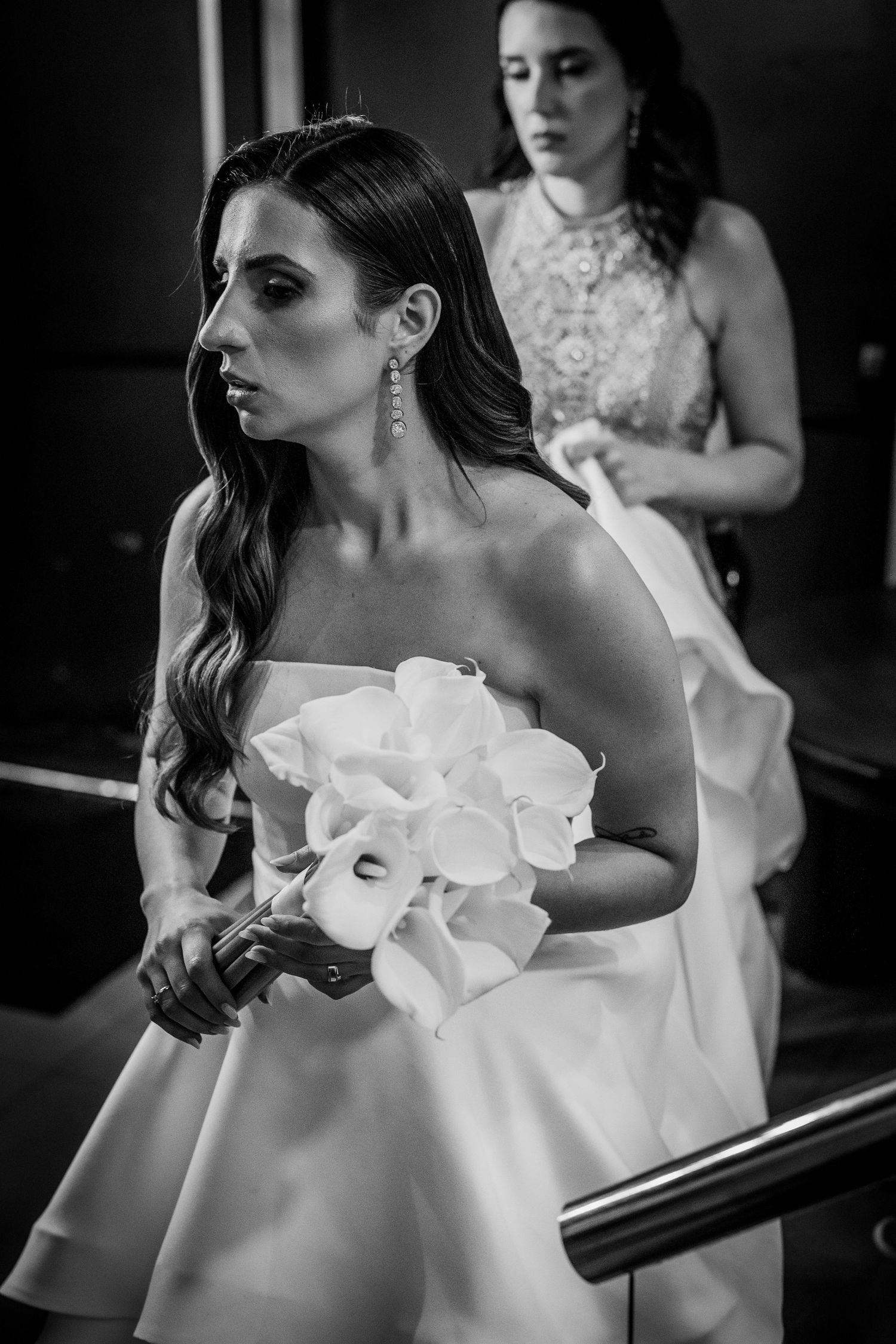 Black and white photo of a bride holding a white calla lily bouquet in a strapless gown, with a bridesmaid behind her.