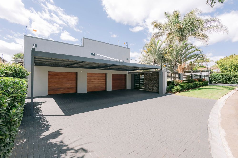 Modern home exterior featuring a triple garage with wooden doors, carport, palm trees, and manicured hedges on a paved driveway.