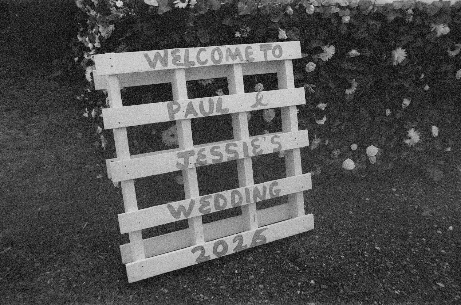 A rustic white wooden pallet wedding welcome sign for Paul and Jessie's 2026 wedding, photographed in black and white.