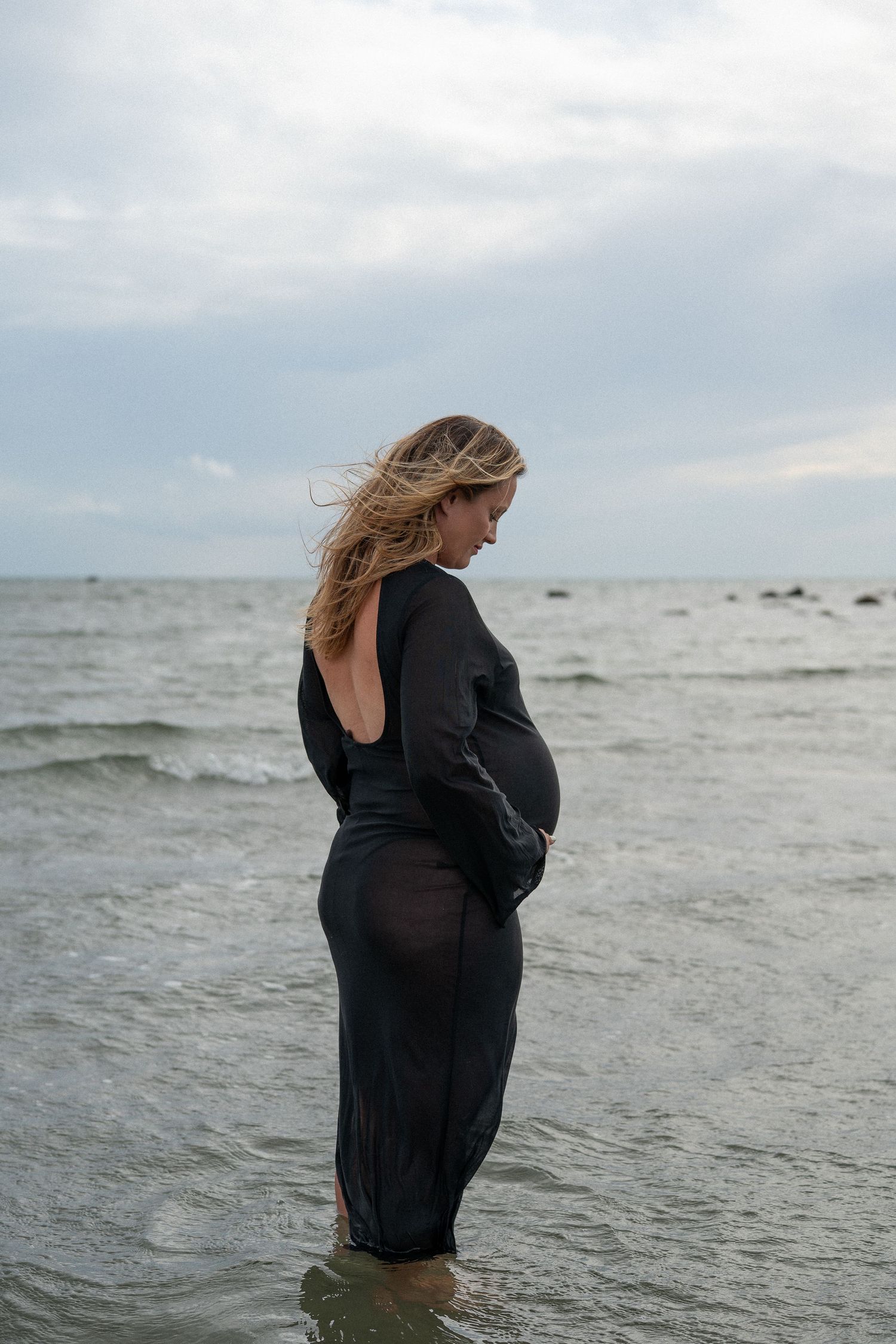 Pregnant woman in black open-back dress standing in ocean waves.