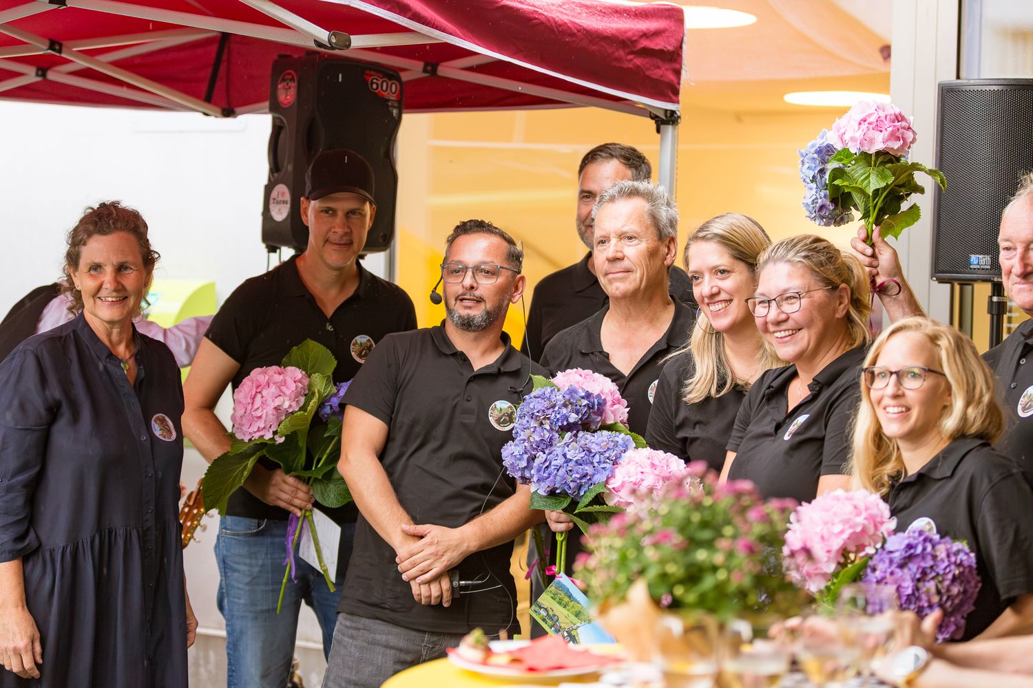 Group of people in black shirts celebrating with colorful hydrangea bouquets at an outdoor event under a red canopy.