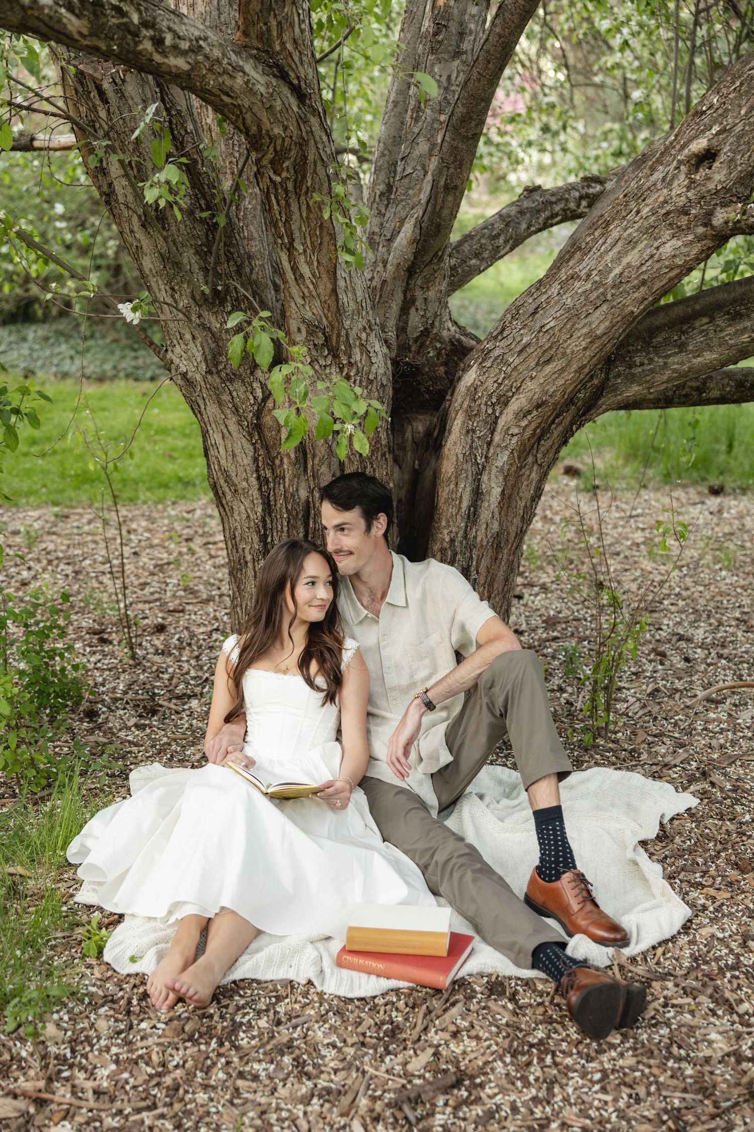 A couple sits under a large tree, woman in white dress reading a book, man in casual attire gazing away, books nearby.