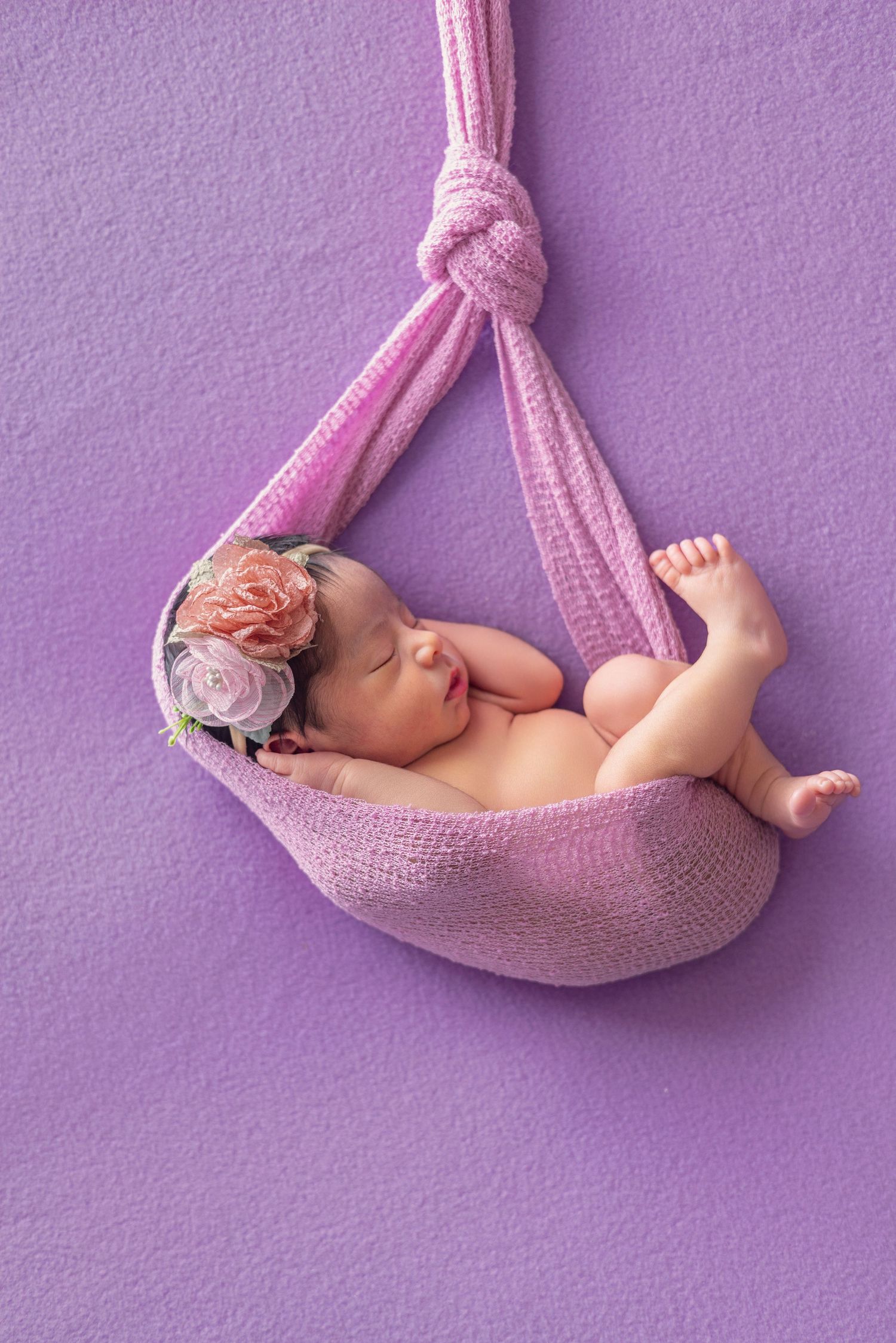 Newborn baby girl sleeping in a pink knit hammock with floral headband on purple background.