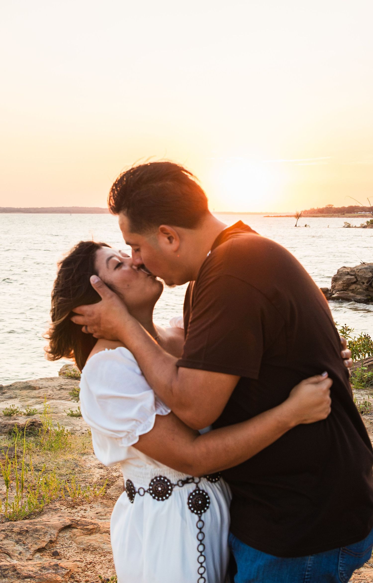 Couple sharing a romantic kiss at sunset by the ocean, woman in white dress, man in black shirt.
