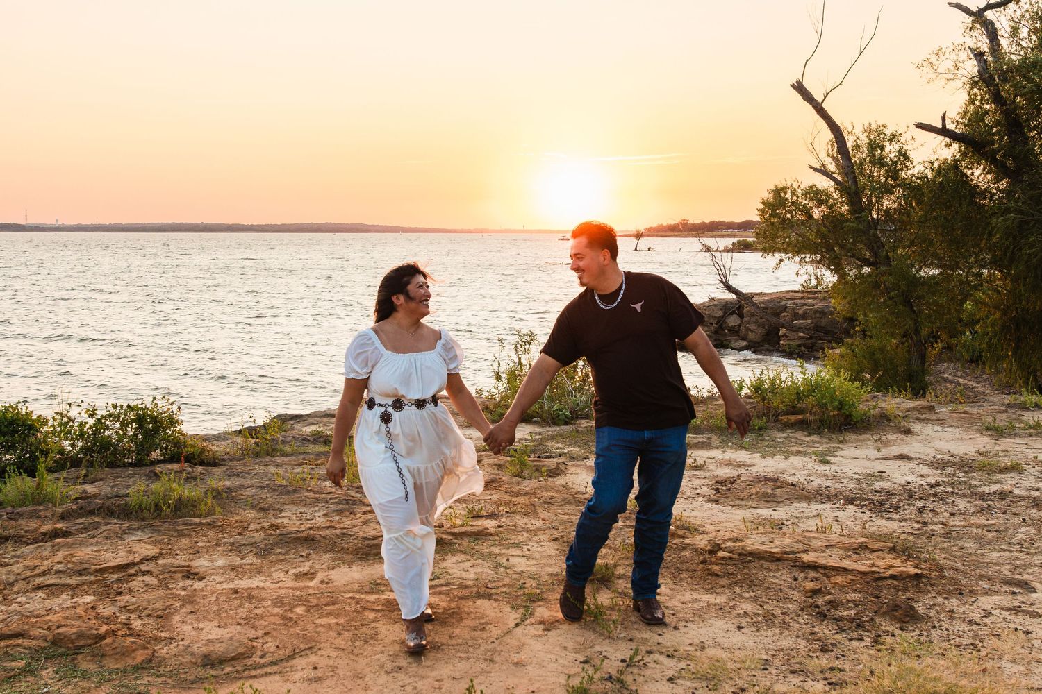 Couple holding hands walking along rocky lakeside cliff at golden sunset, woman in white dress, man in black shirt.
