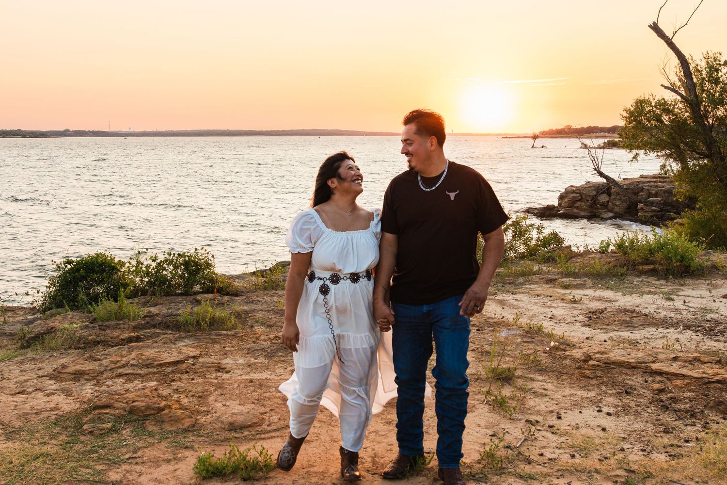 Couple holding hands smiling at sunset by a lake, woman in white outfit, man in dark shirt and jeans.