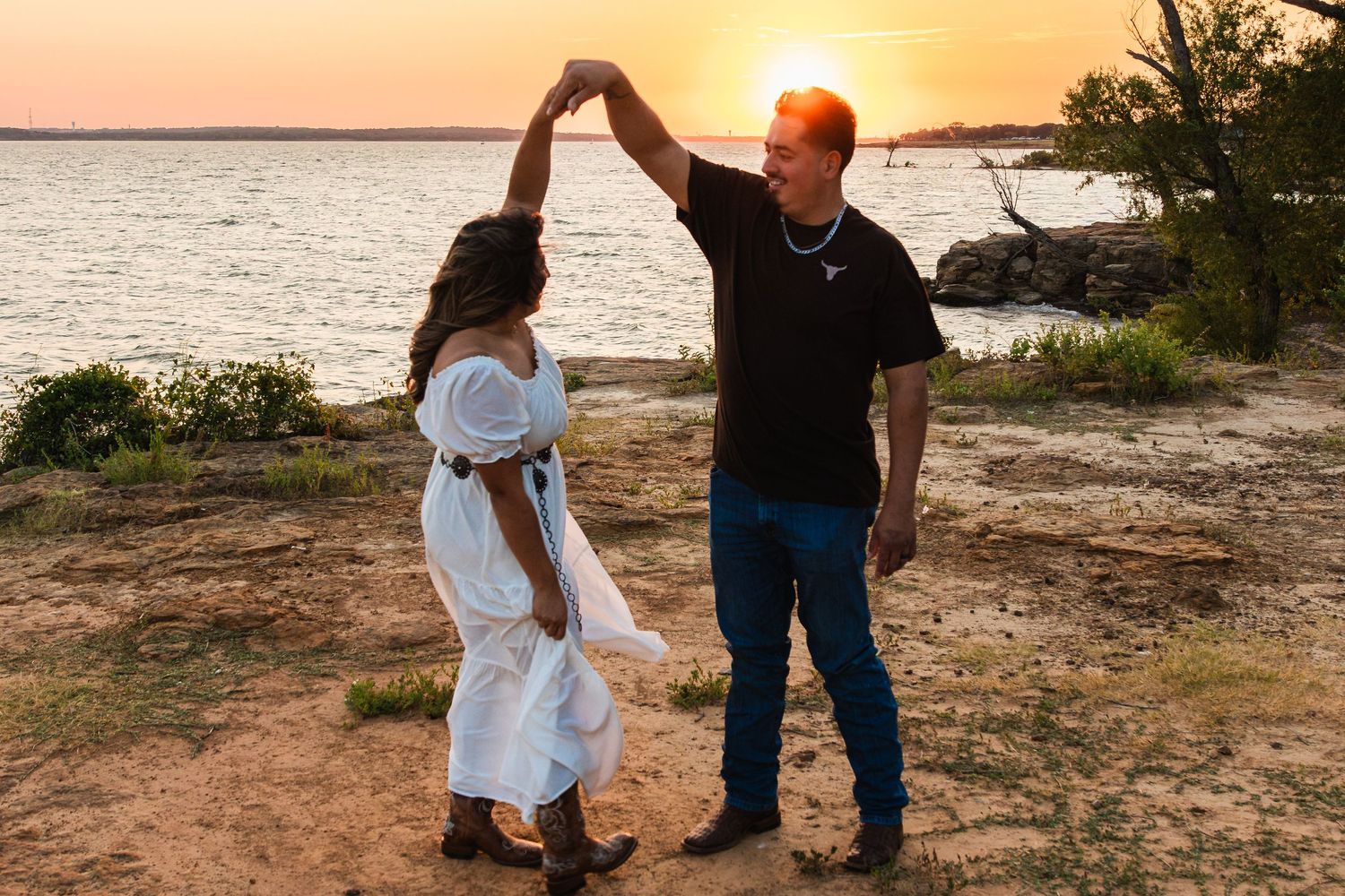 Couple dancing at sunset by the water, woman in white dress, man in black shirt and jeans, golden hour glow.