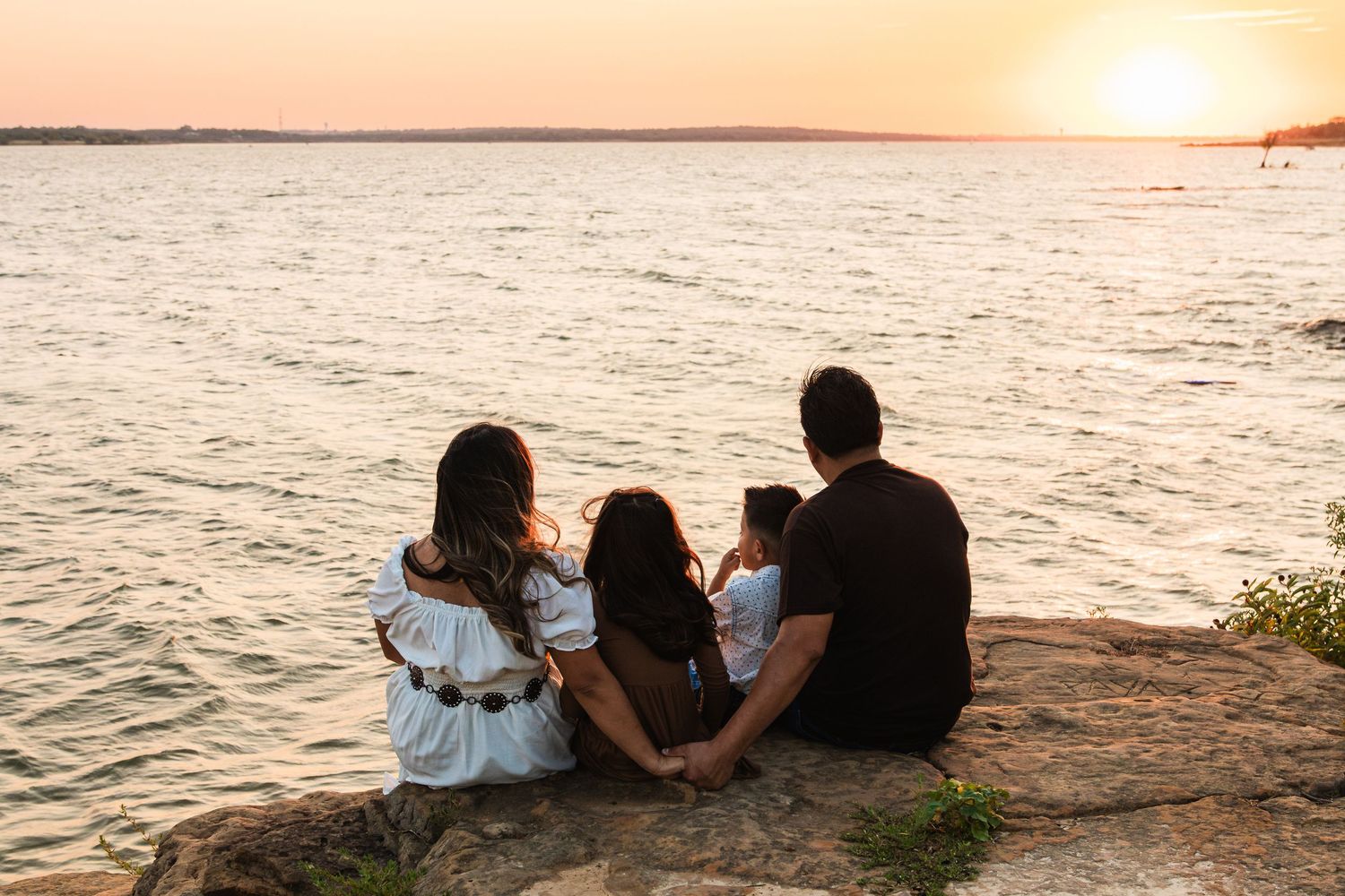 Family of four sitting on rocky shore watching golden sunset over calm water.