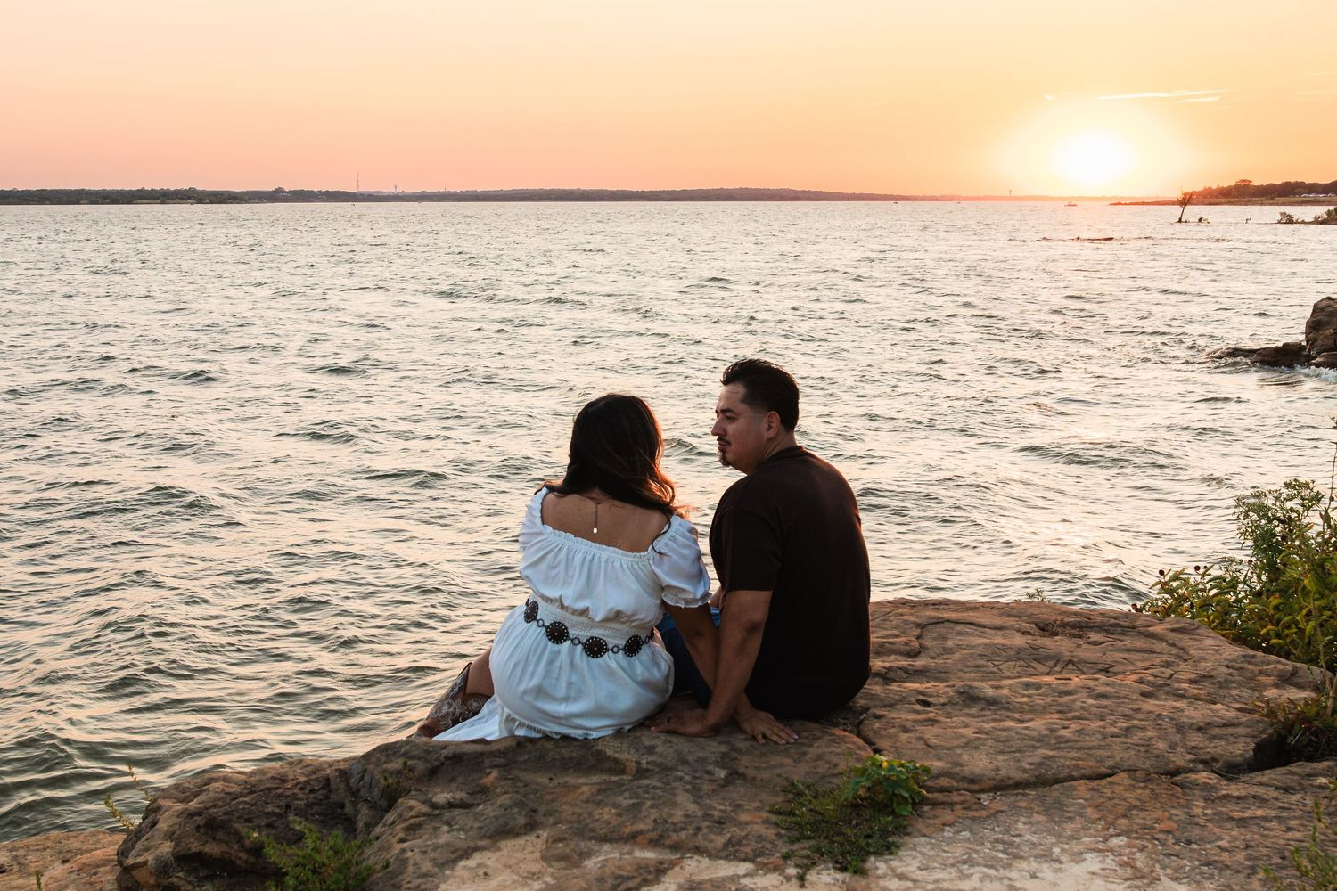 Couple sitting on rocky shore watching golden sunset over calm water, romantic outdoor scene.
