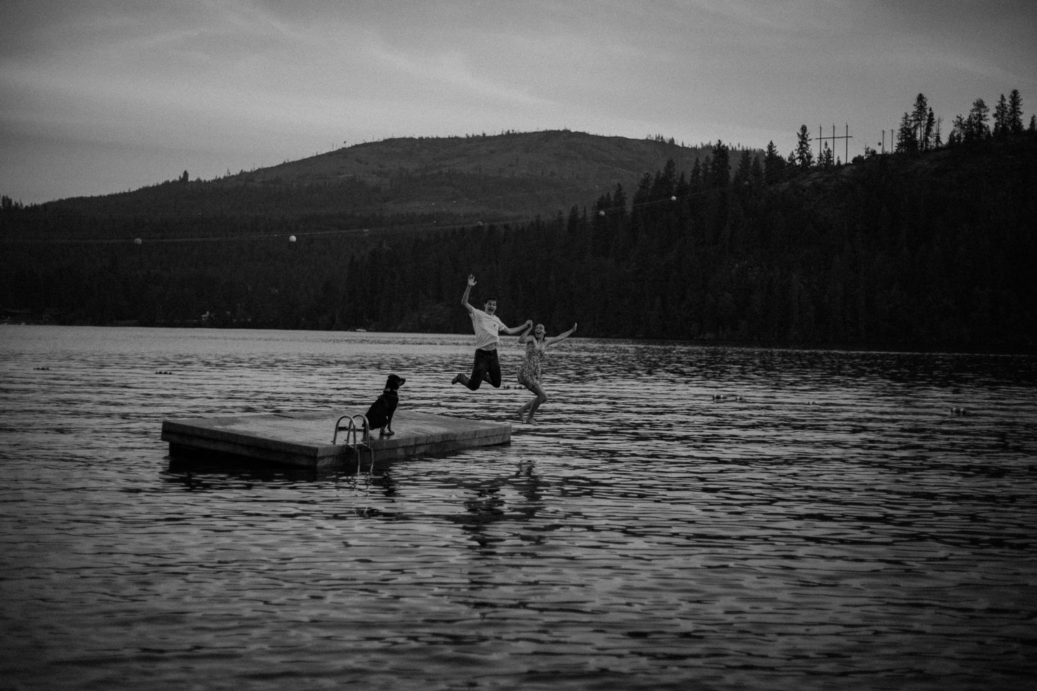 Black and white photo of a person jumping off a lake dock at dusk with hills and trees in background.